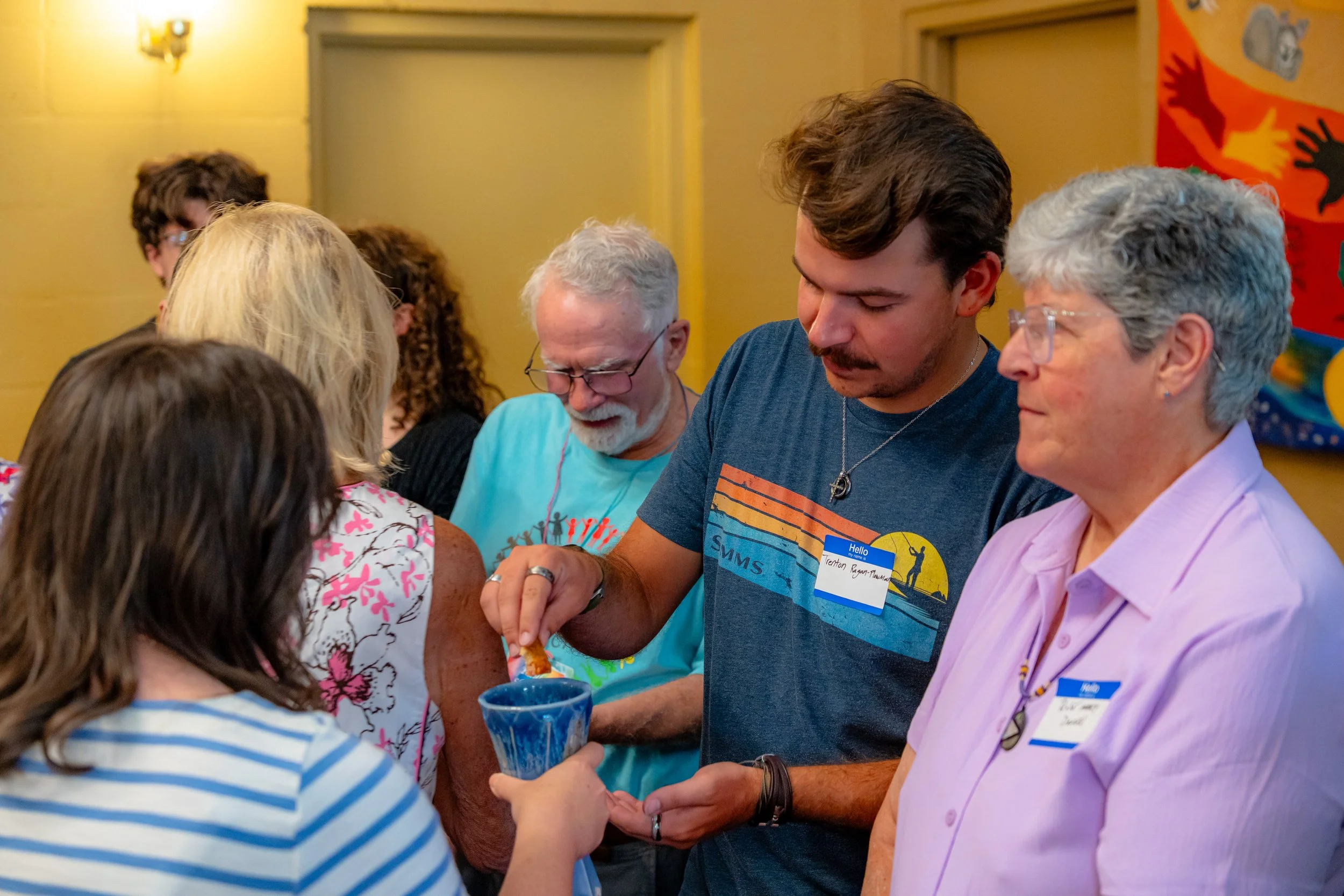 A group of people gathered closely at an indoor event, engaged in a conversation or activity. The scene includes a woman with gray hair and glasses, an elderly man with glasses, a young man with dark hair, and a woman with short gray hair, all appear