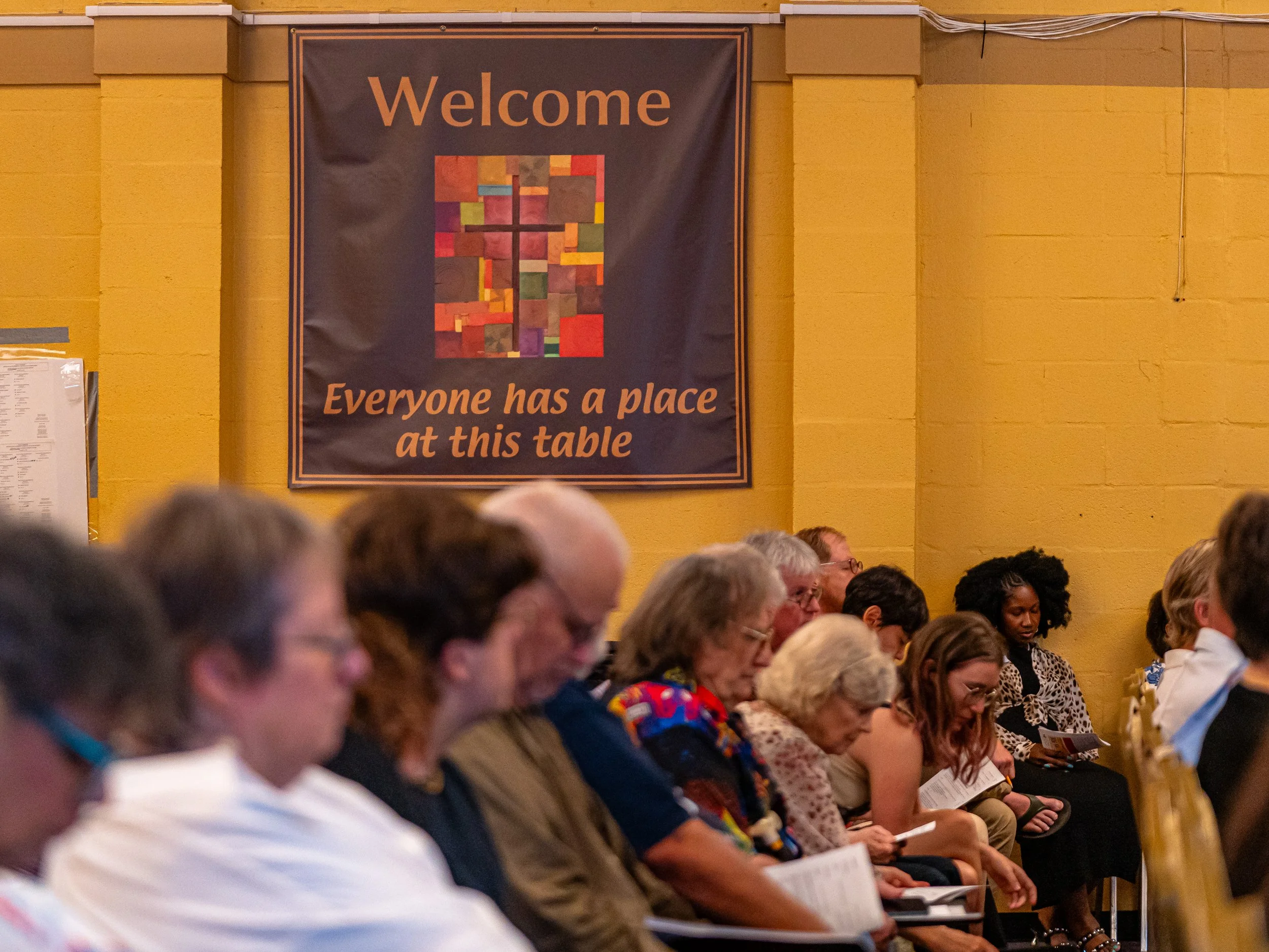 People sitting in an audience in a large room with a yellow wall, listening to a speaker or presentation, with a banner that says "Welcome" and "Everyone has a place at this table" hanging on the wall.