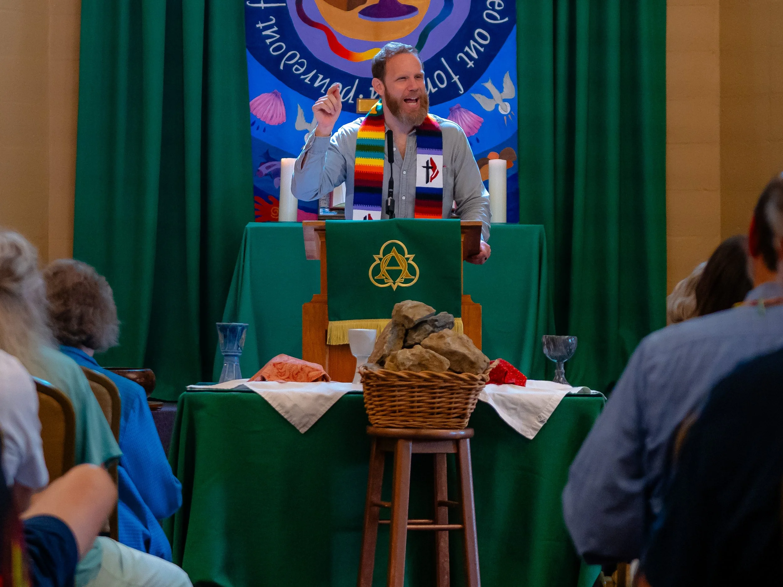 A man with a beard and mustache standing at a podium, speaking or giving a presentation with a colorful backdrop behind him, decorated with symbols, paintings, or logos, and green curtains on either side.