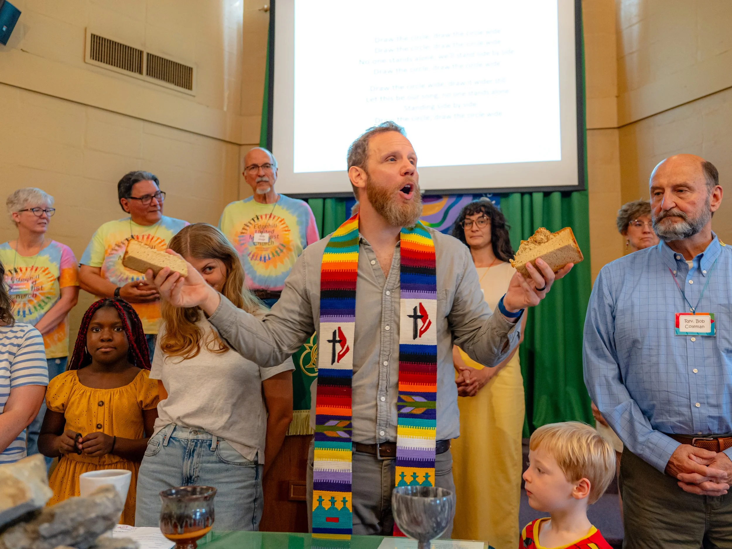 A man with a beard is holding a slice of cake and appears to be singing or speaking, wearing a colorful striped stole around his neck. Other people are gathered around, some wearing tie-dye shirts, and there is a large screen in the background with t
