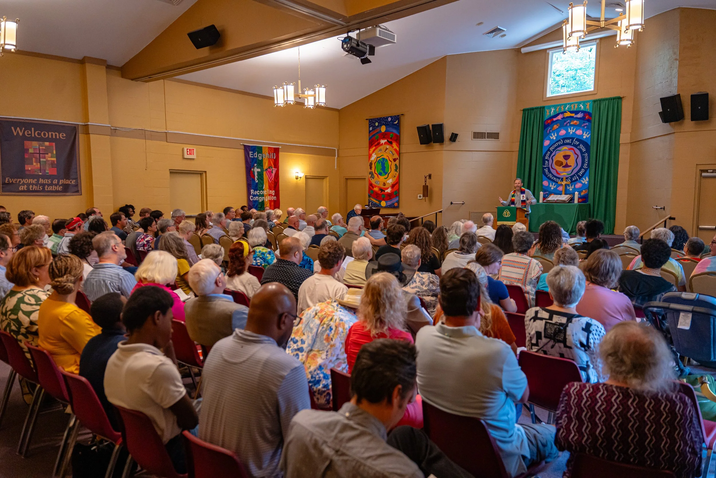 A large group of people attending a church service or community gathering in a synagogue and listening to a speaker at a podium on stage. The room has beige walls, colorful banners, and a green curtain behind the speaker.