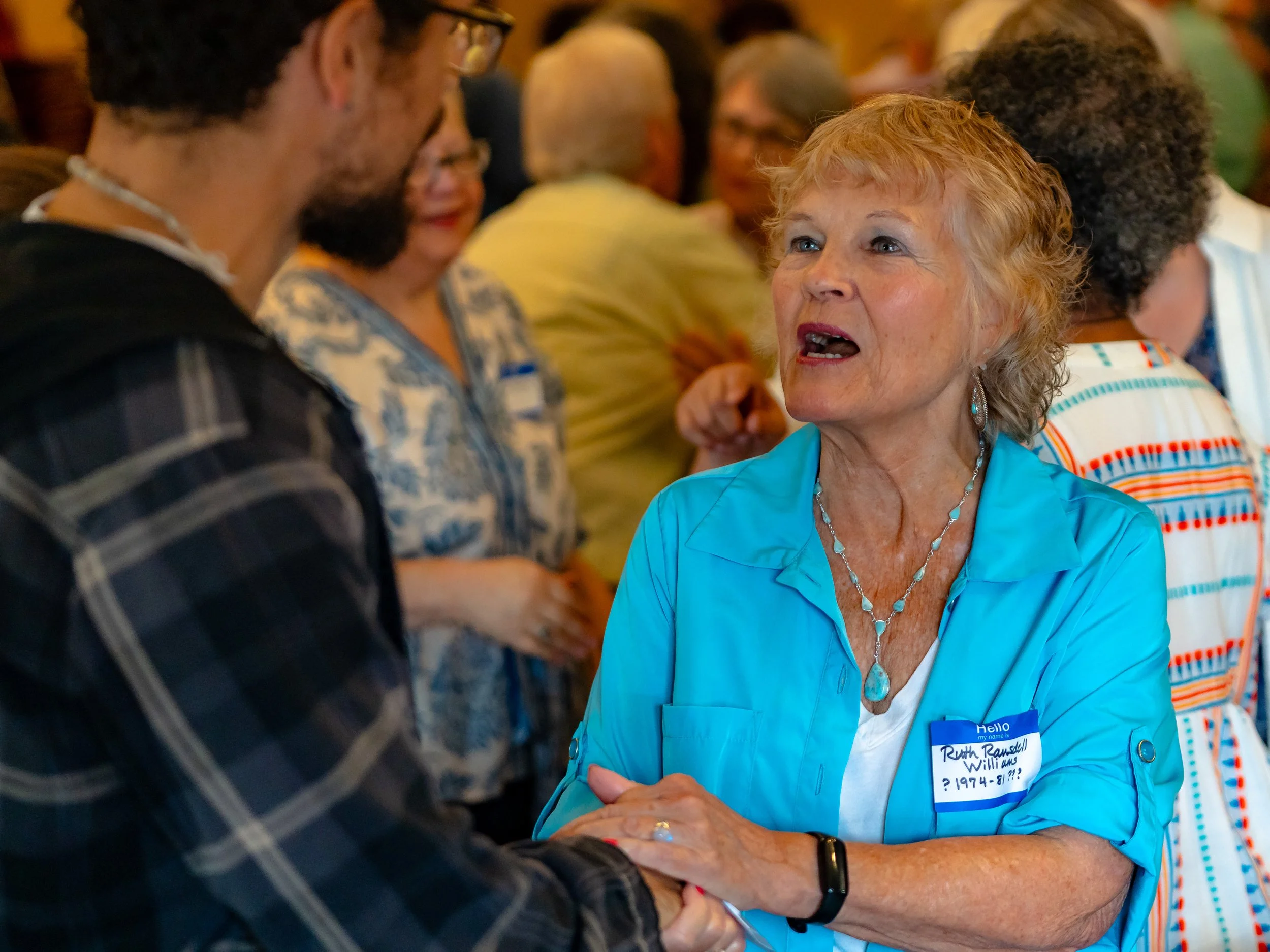 A woman with curly blonde hair wearing a blue jacket and name tag talking to a person with glasses and dark hair in a busy indoor setting.