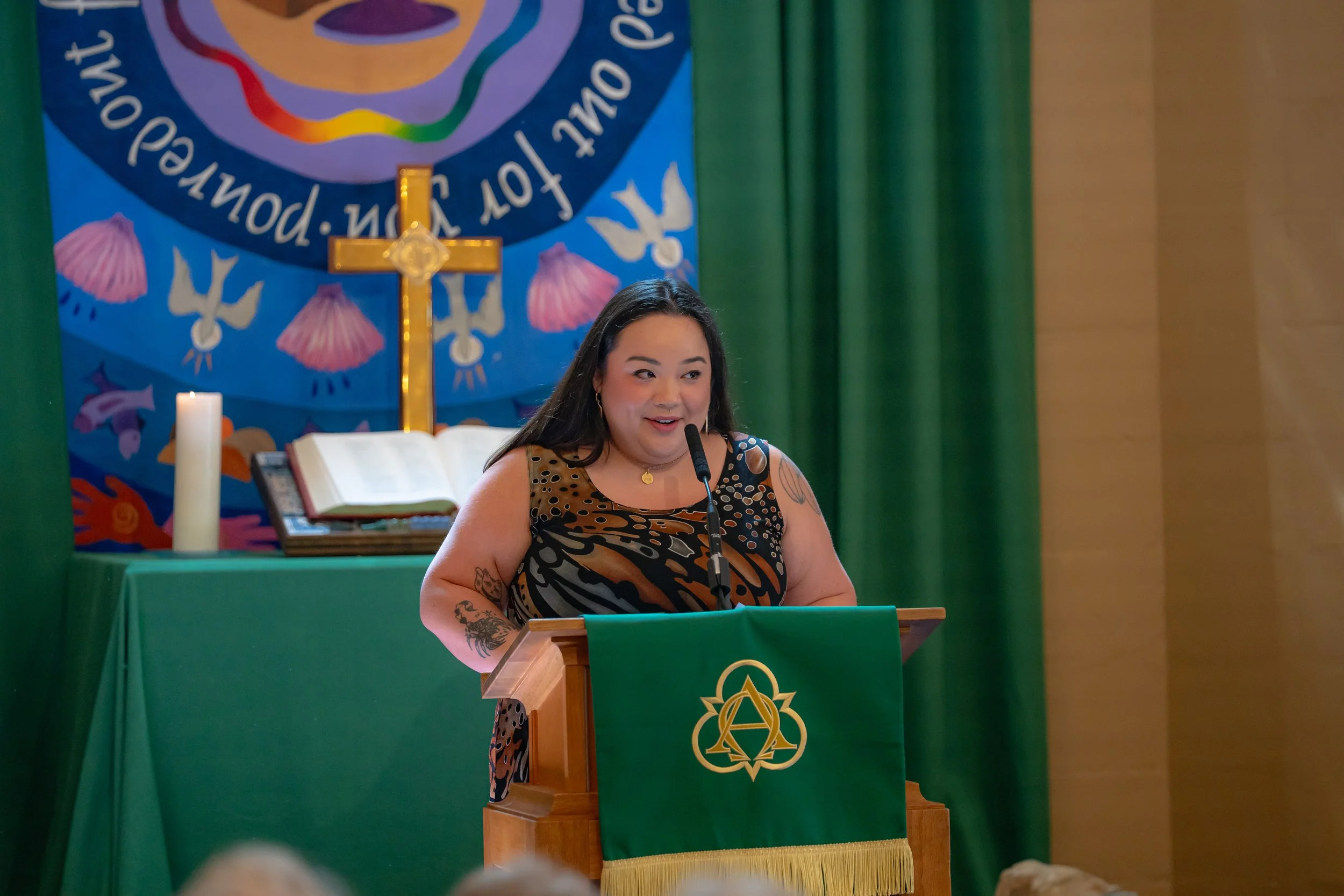 A woman with long dark hair, wearing earrings and tattoos on her arms, stands at a podium with a microphone, speaking. Behind her, there is a colorful banner with religious symbols, a cross, and a Bible open on a green table. The setting appears to b