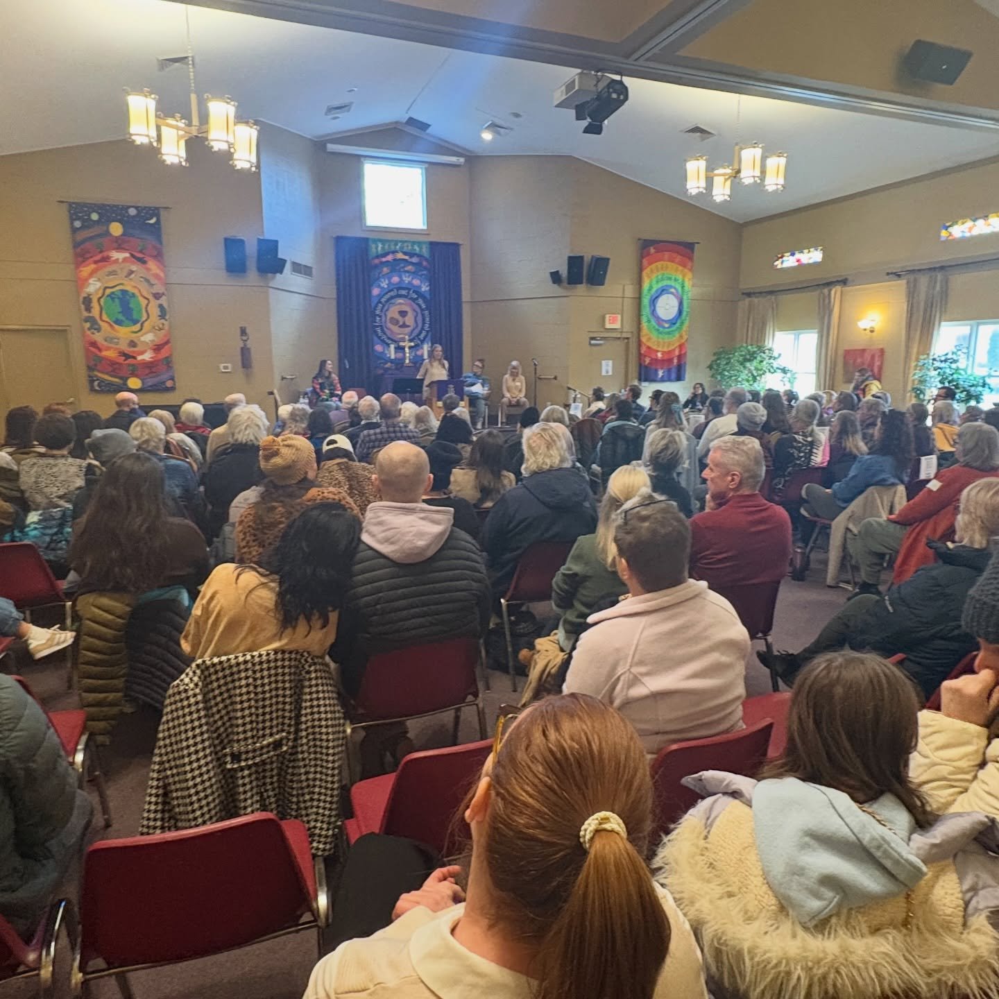 A large gathering of people seated in an auditorium listening to a panel of speakers at the front of the room, decorated with colorful banners and stained glass windows.
