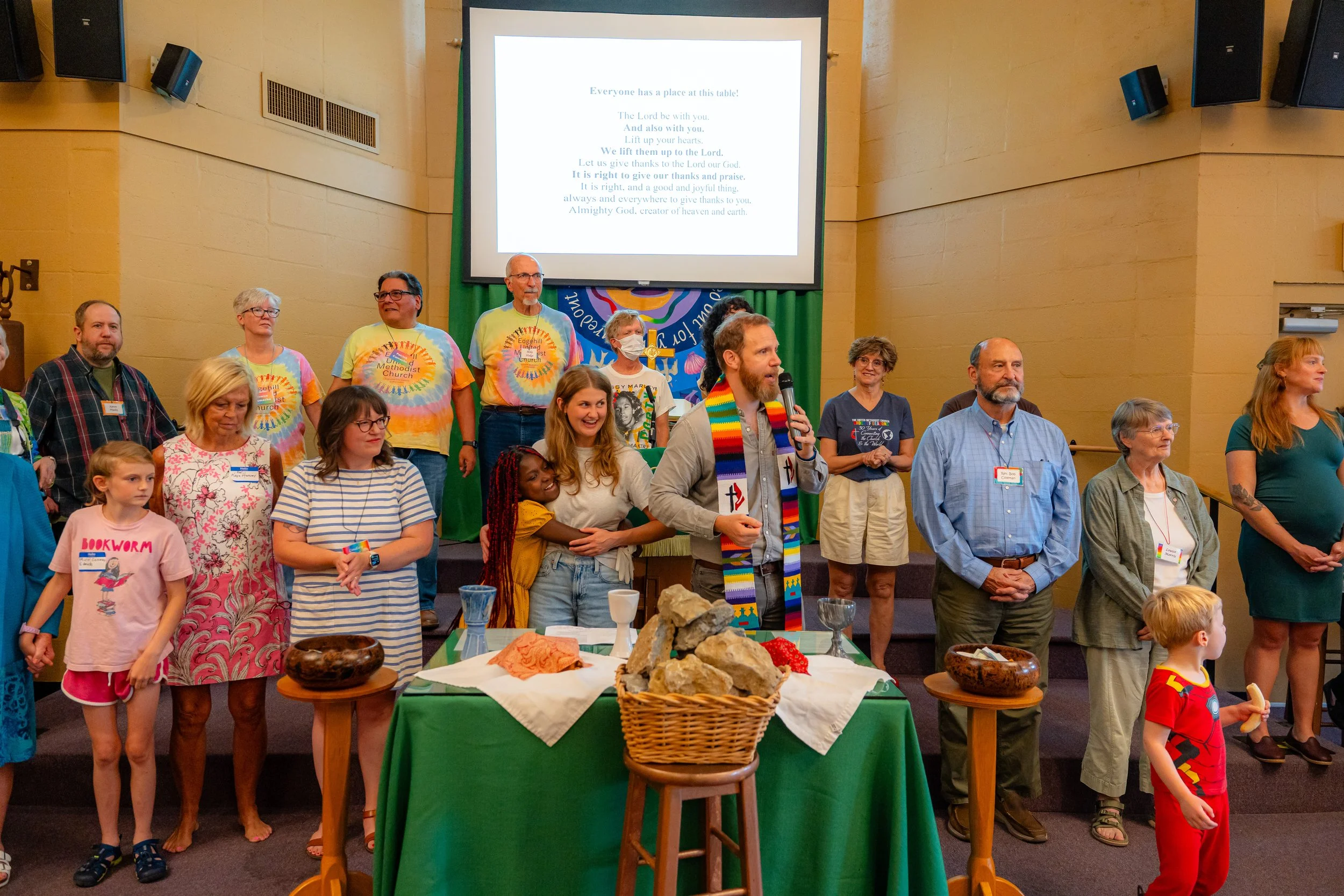 A diverse group of people, including children and adults, gathered in a church or community hall, standing behind a table with rocks and textiles. There is a large screen displaying a religious message about giving thanks to the Lord. The group appea