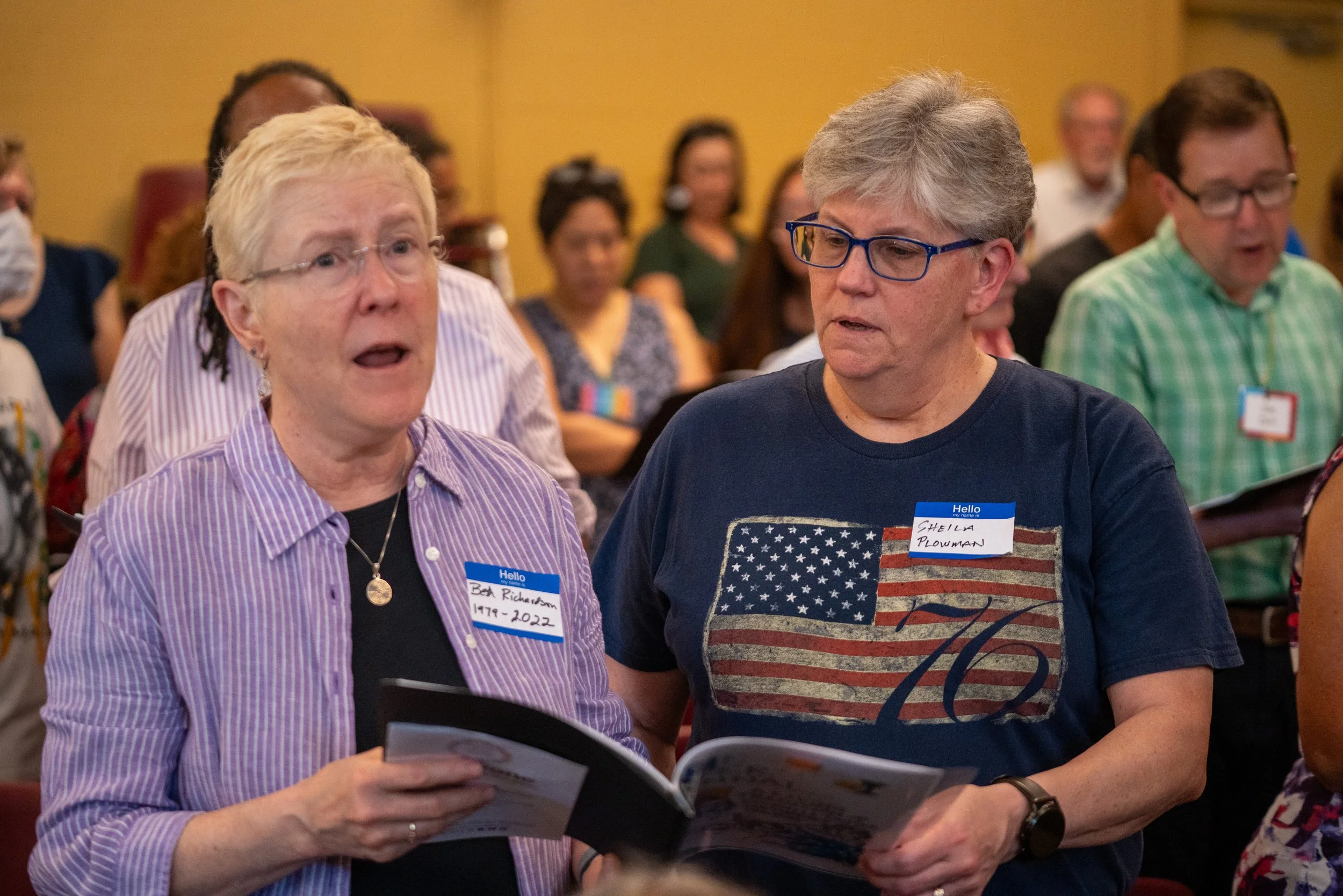 Two women in a crowded room looking at a brochure or program, engaging in conversation. The woman on the left has short, light-colored hair, glasses, and a purple striped shirt. The woman on the right has short gray hair, glasses, and a shirt with an