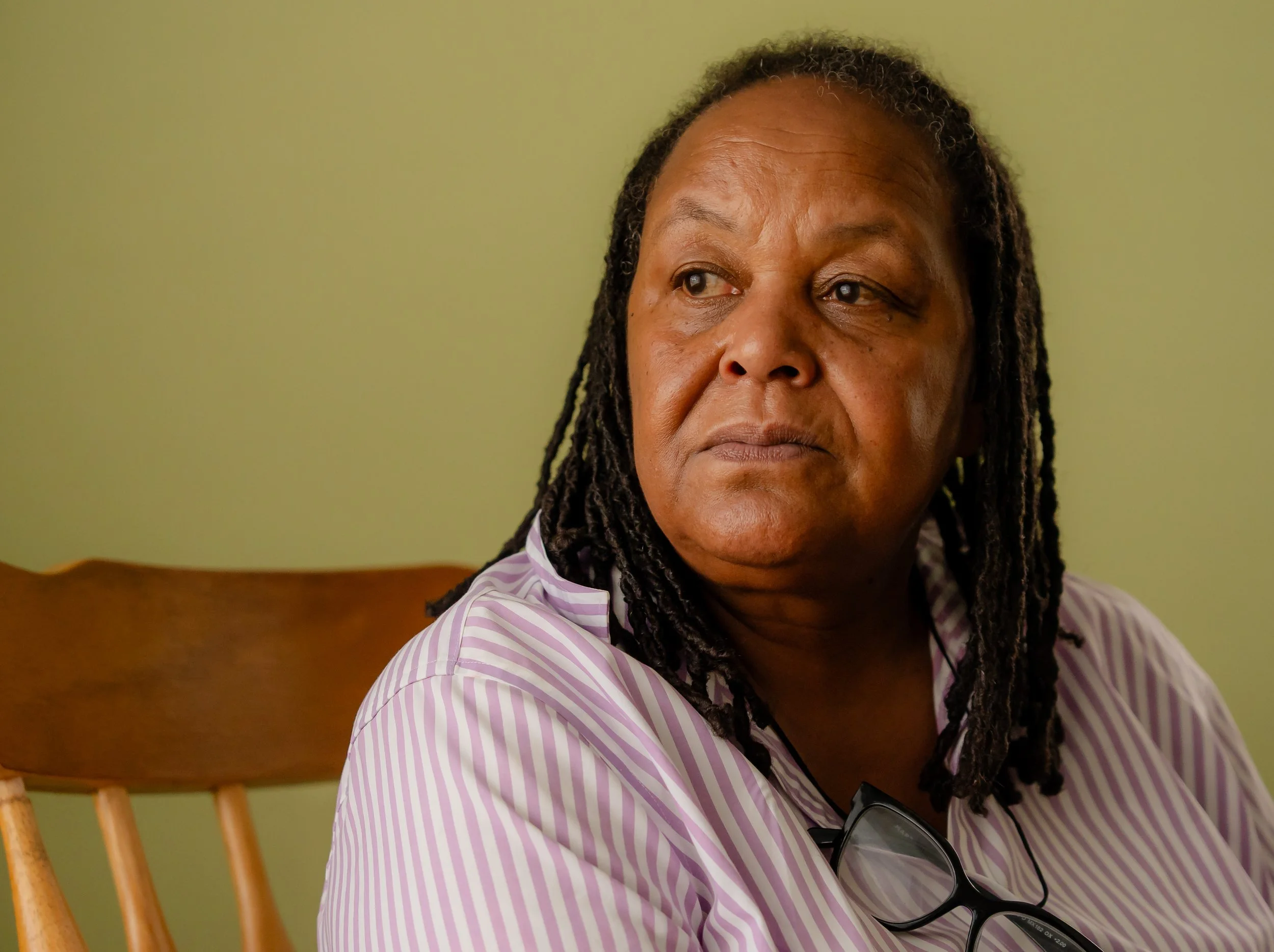 Close-up of a woman with dreadlocks wearing a pink and white striped shirt, sitting on a wooden chair, with glasses hanging on her shirt, and a neutral expression as she looks to the side.