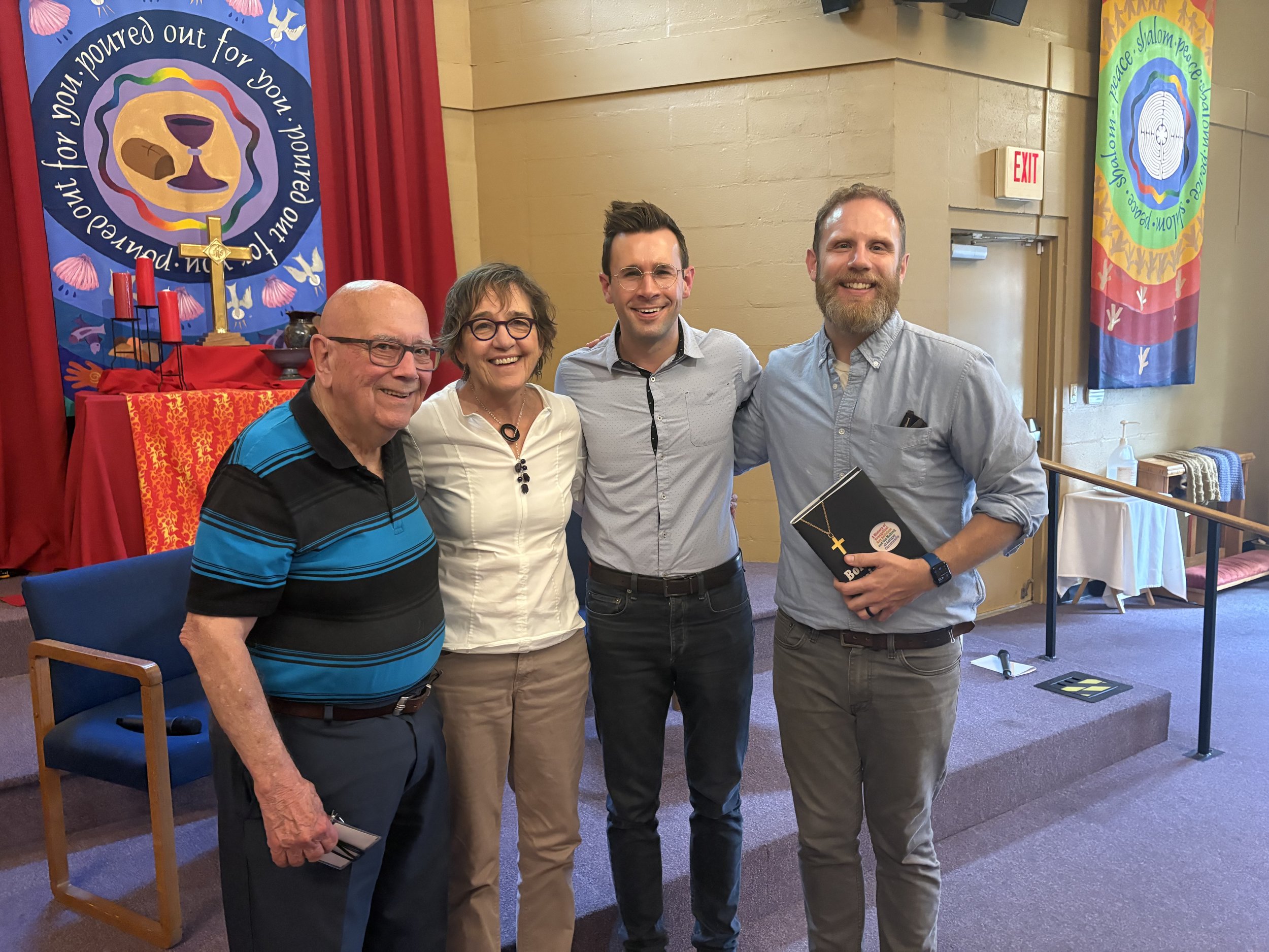 A group of four people standing together, smiling, in front of colorful banners with religious symbols, during an event or celebration.
