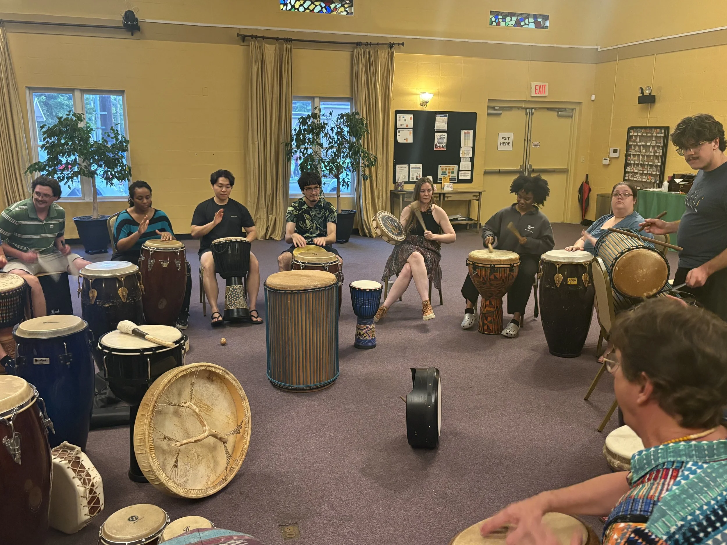 A group of eight people sitting in a circle playing various types of drums in an indoor space with yellow walls, windows, and plants.