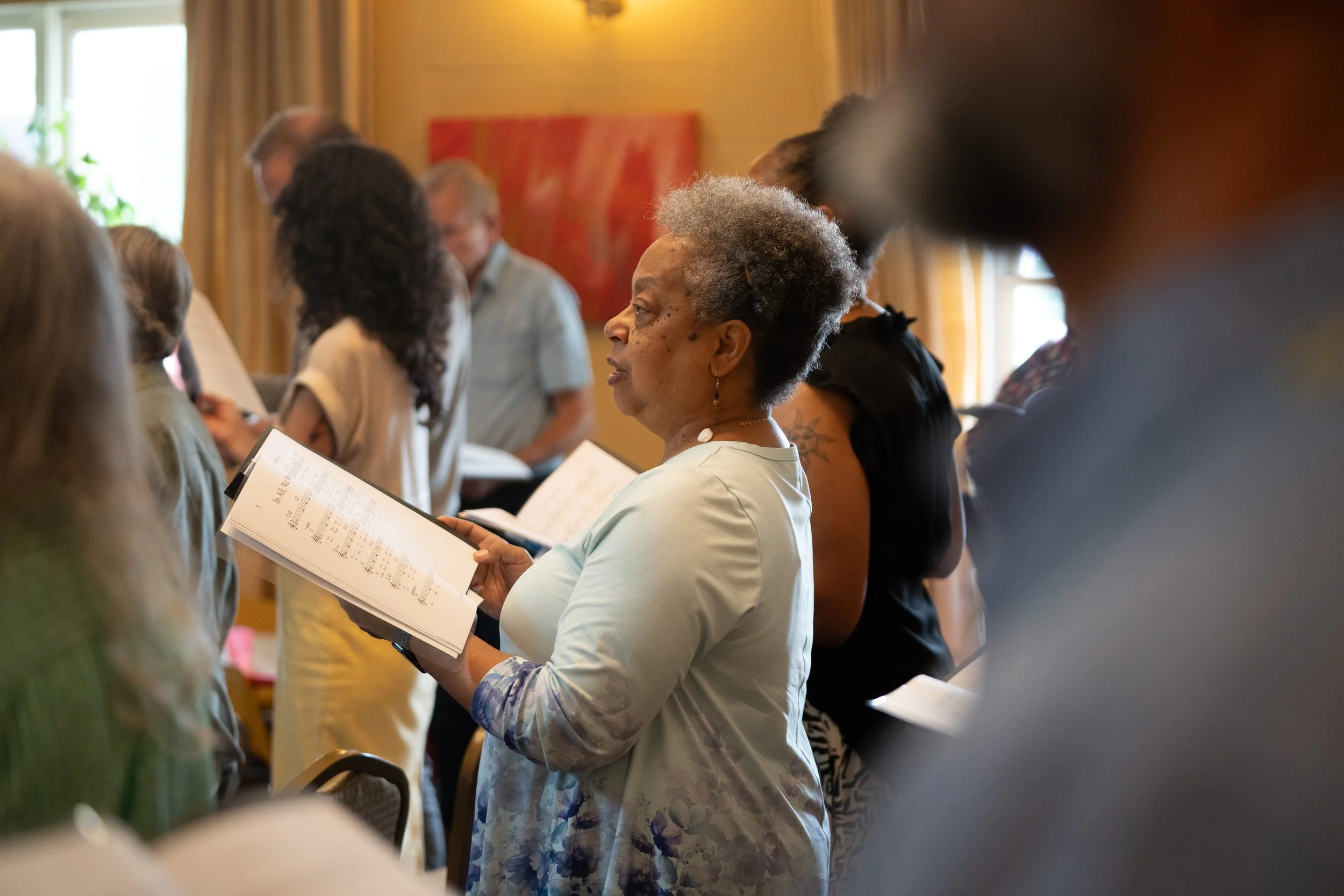 A group of people standing in a room, holding sheet music, likely participating in a choir or singing practice session.