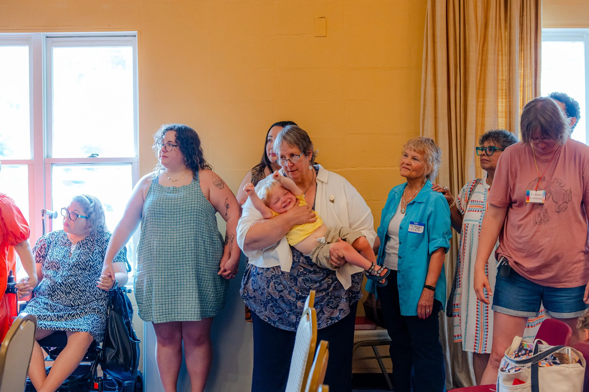 Group of women, some with glasses, standing in a room with yellow walls and large windows, attending an event.