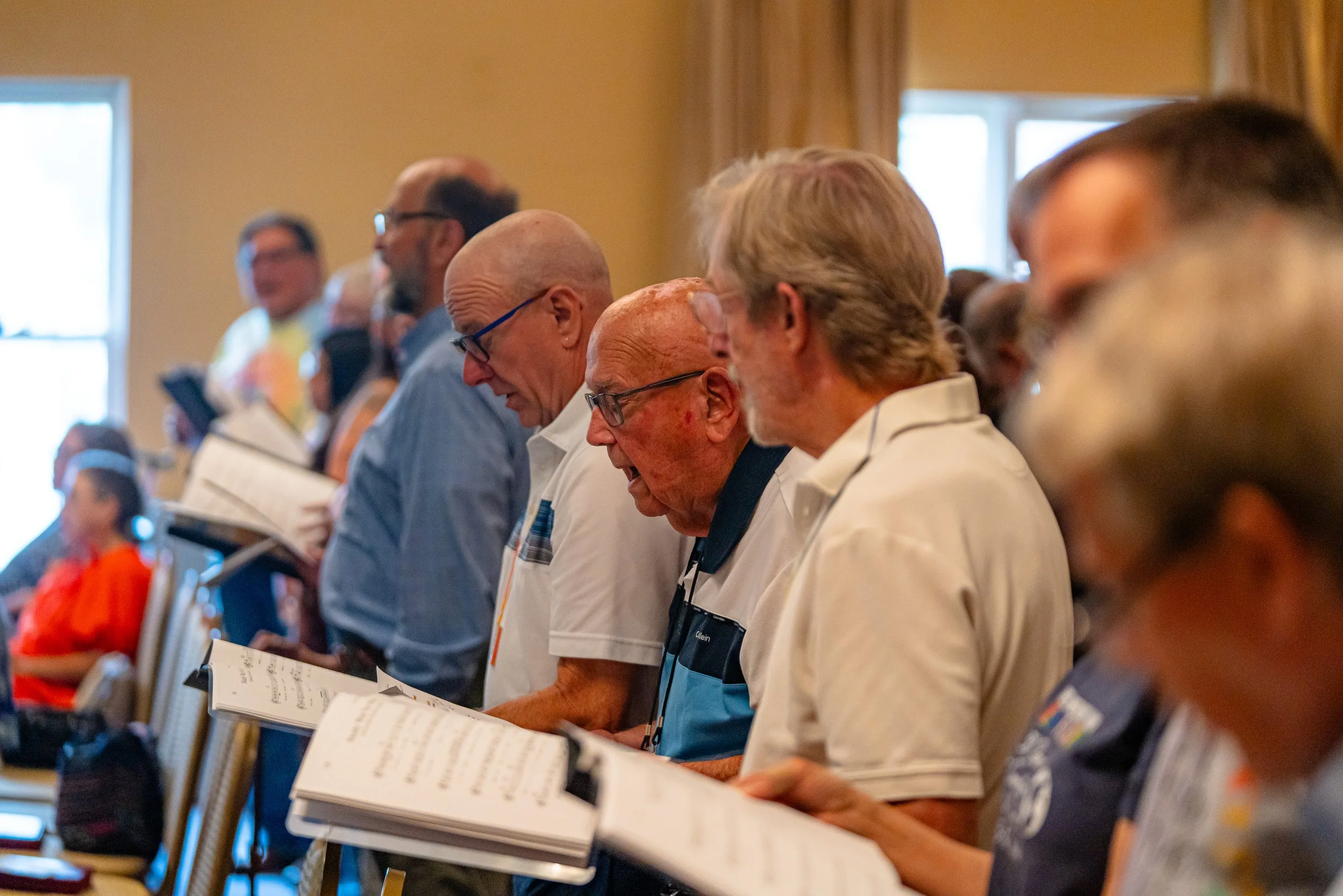 A diverse group of people, mostly elderly, standing in a row inside a room with yellow walls and large windows, looking at open hymnals or prayer books.