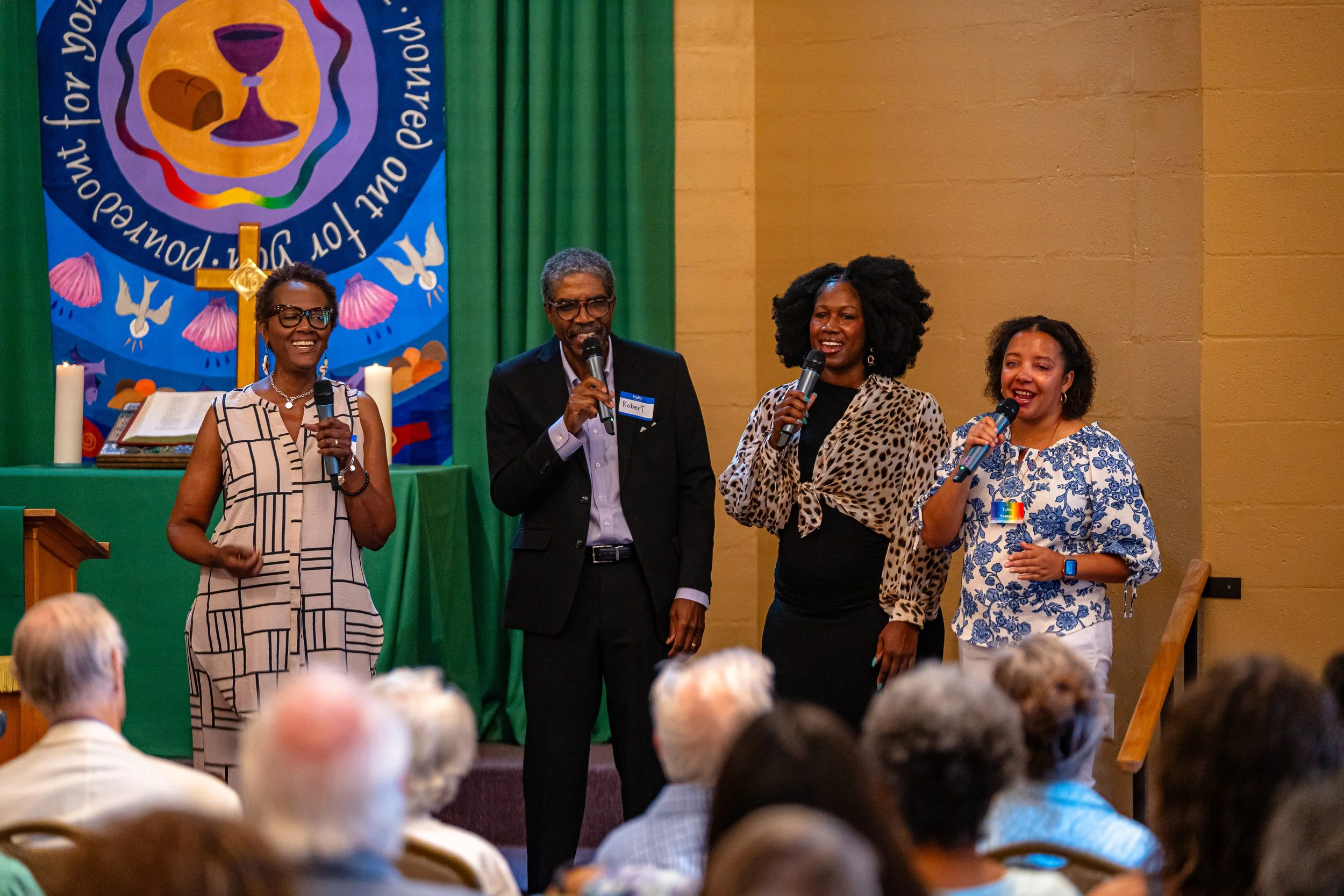 Four people standing on a stage, holding microphones, attending a formal event with an audience. Behind them is a banner with religious symbols and Dove images. The audience, mostly elderly, watches the scene.