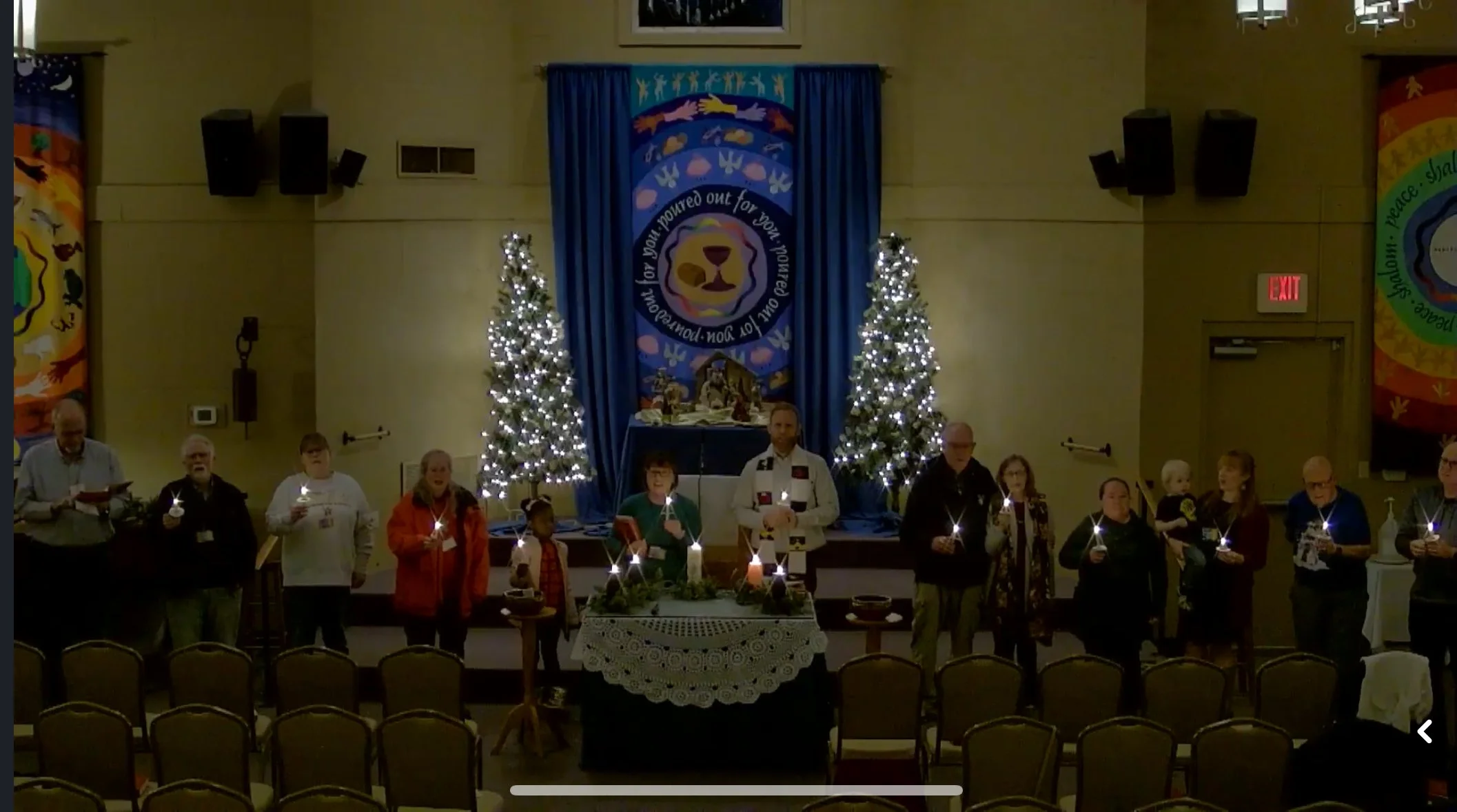 People standing on a stage holding candles, in front of holiday decorations including two decorated Christmas trees, with a large blue banner featuring a lion and a circular message in the background.