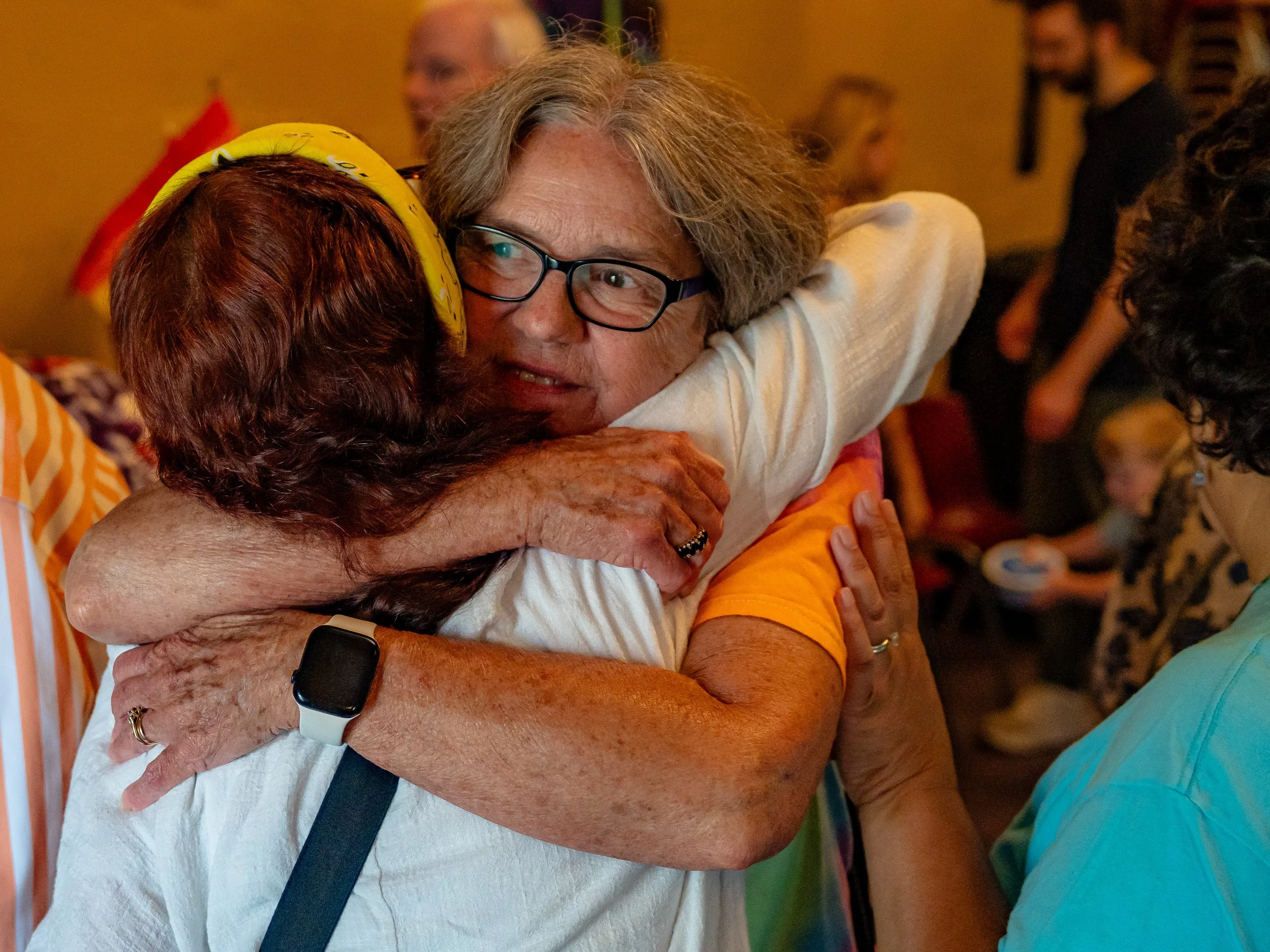 Two women hugging at an indoor gathering, one with red hair wearing a striped shirt and the other with gray hair and glasses wearing a white top.