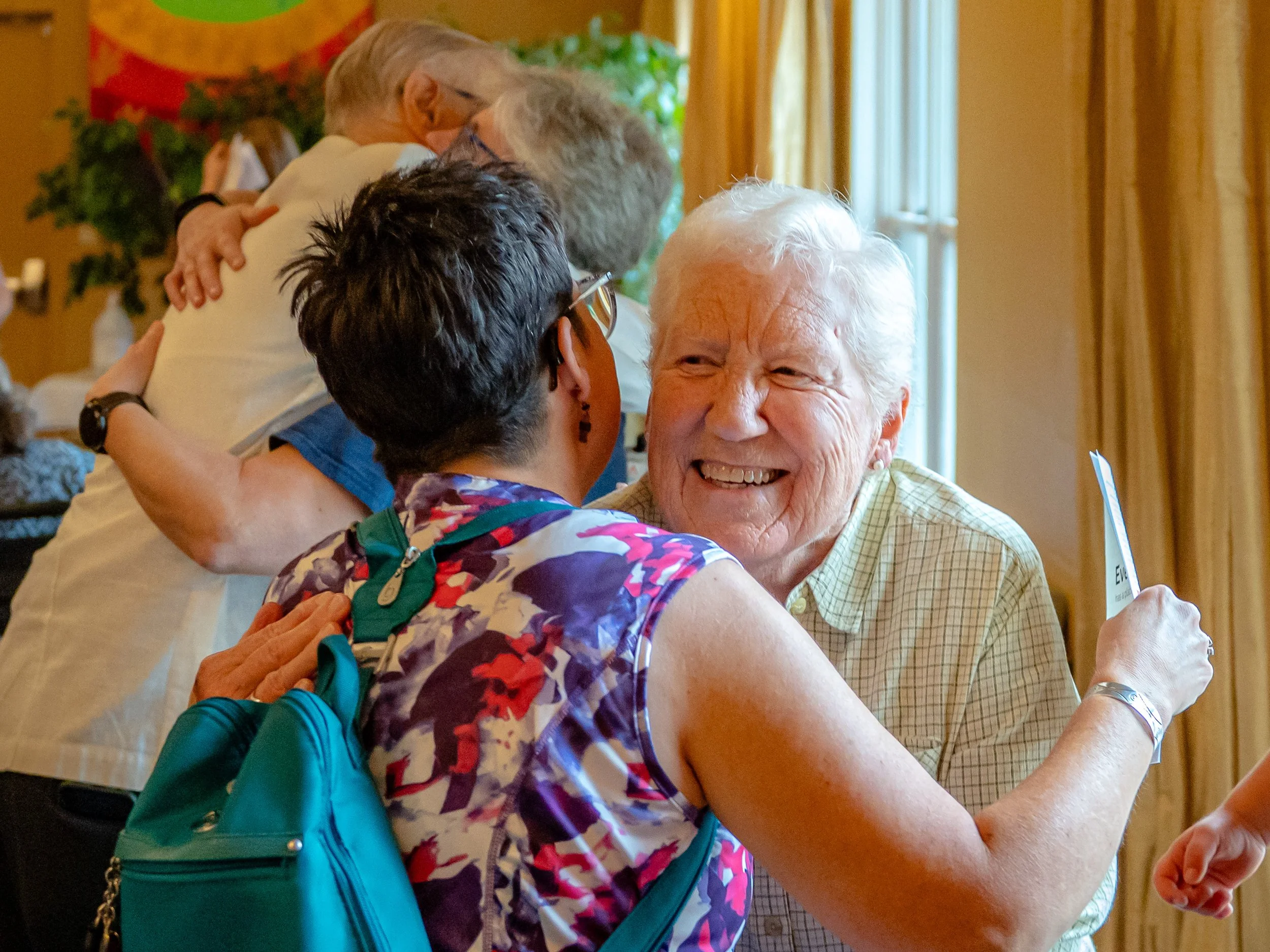 Two elderly women and a middle-aged woman hug each other warmly indoors. They appear to be happy and enjoy each other's company in a brightly lit room with curtains and a poster on the wall.