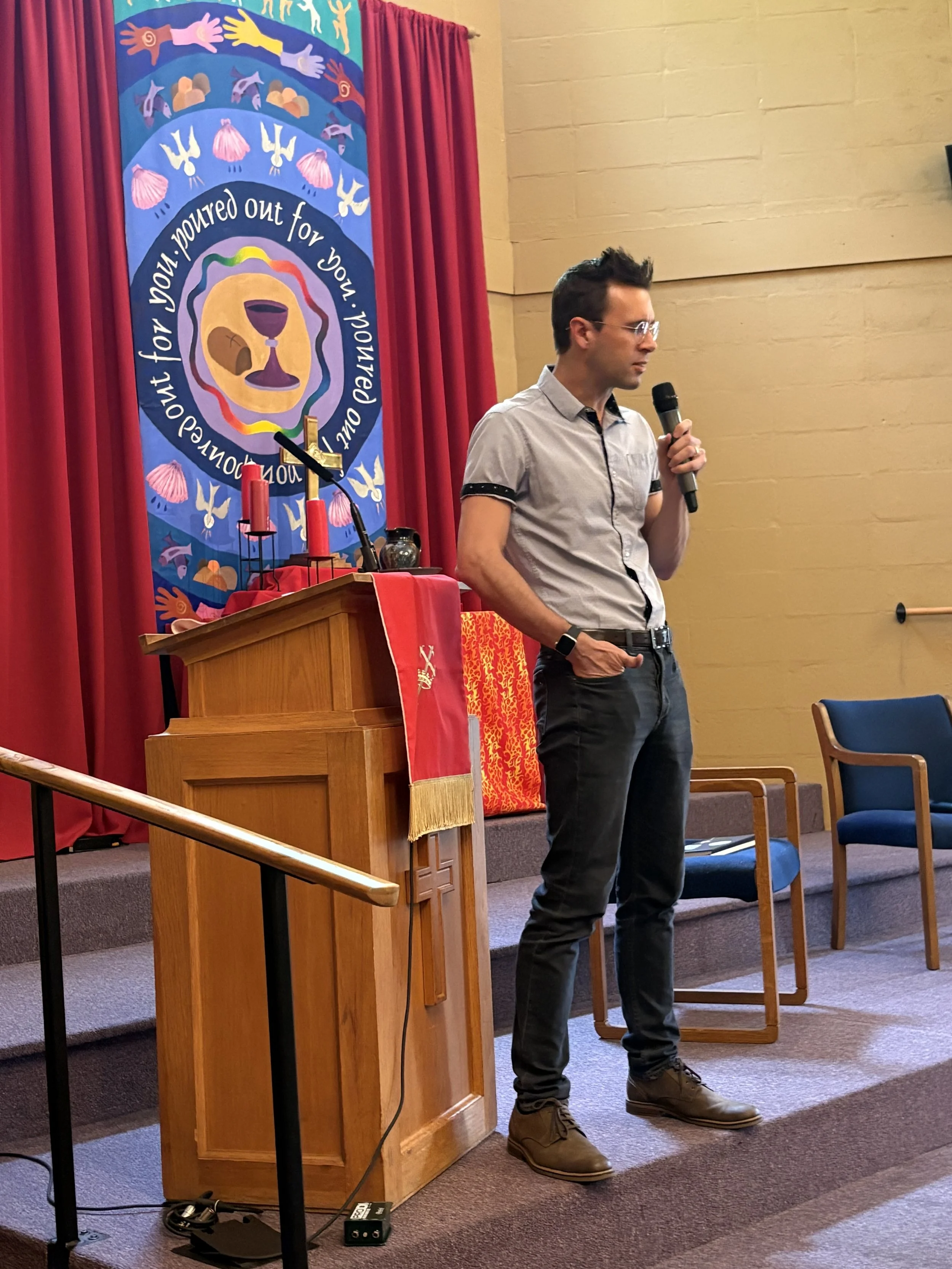 A man in glasses, a light gray short-sleeve button-up shirt with black trim on the sleeves, black pants, and brown shoes is holding a black microphone and standing on a stage next to a wooden pulpit. Behind him is a colorful religious banner with tex