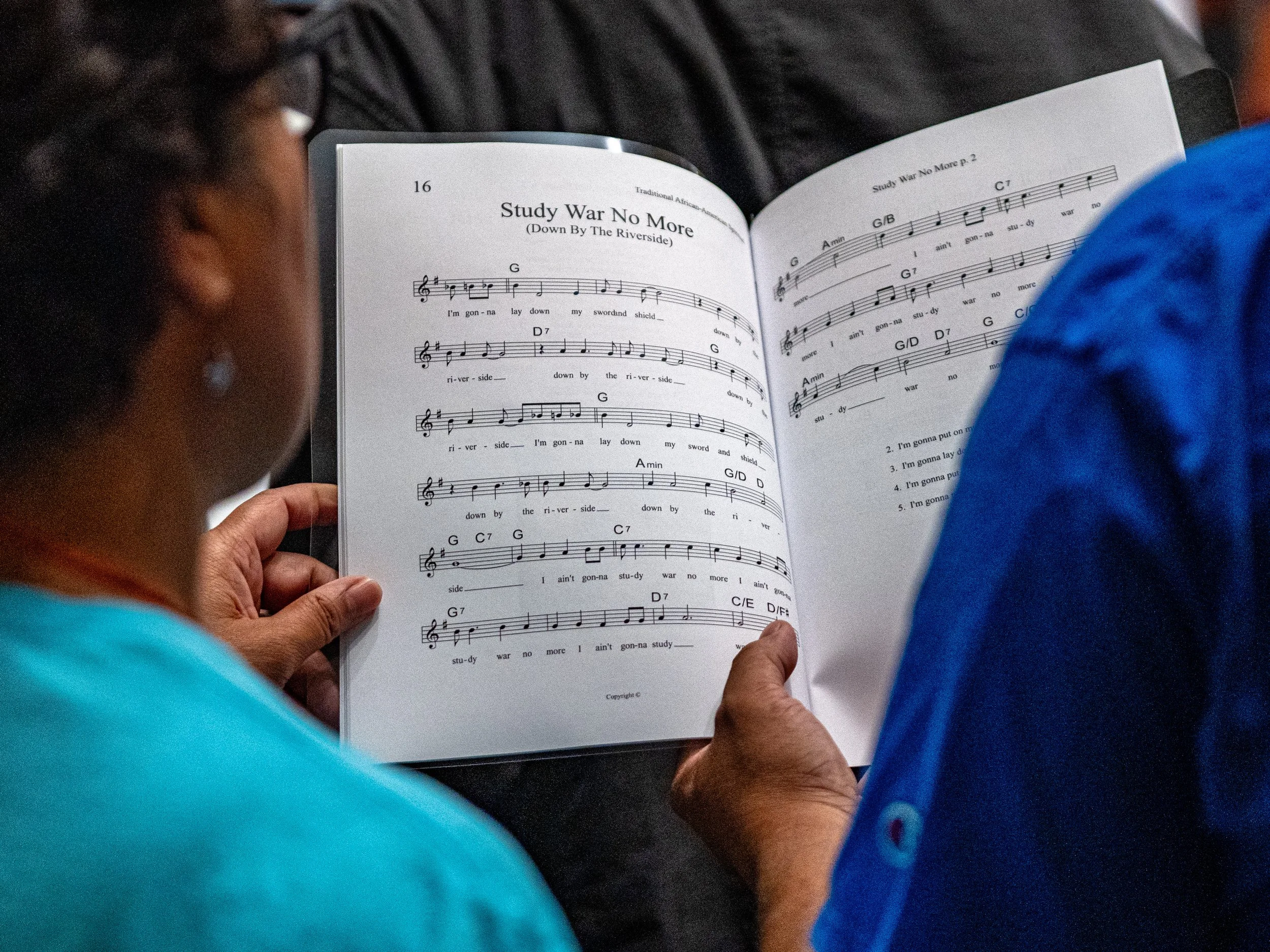 A person holding an open songbook with the title 'Study War No More' and musical notation visible, while another person stands nearby.