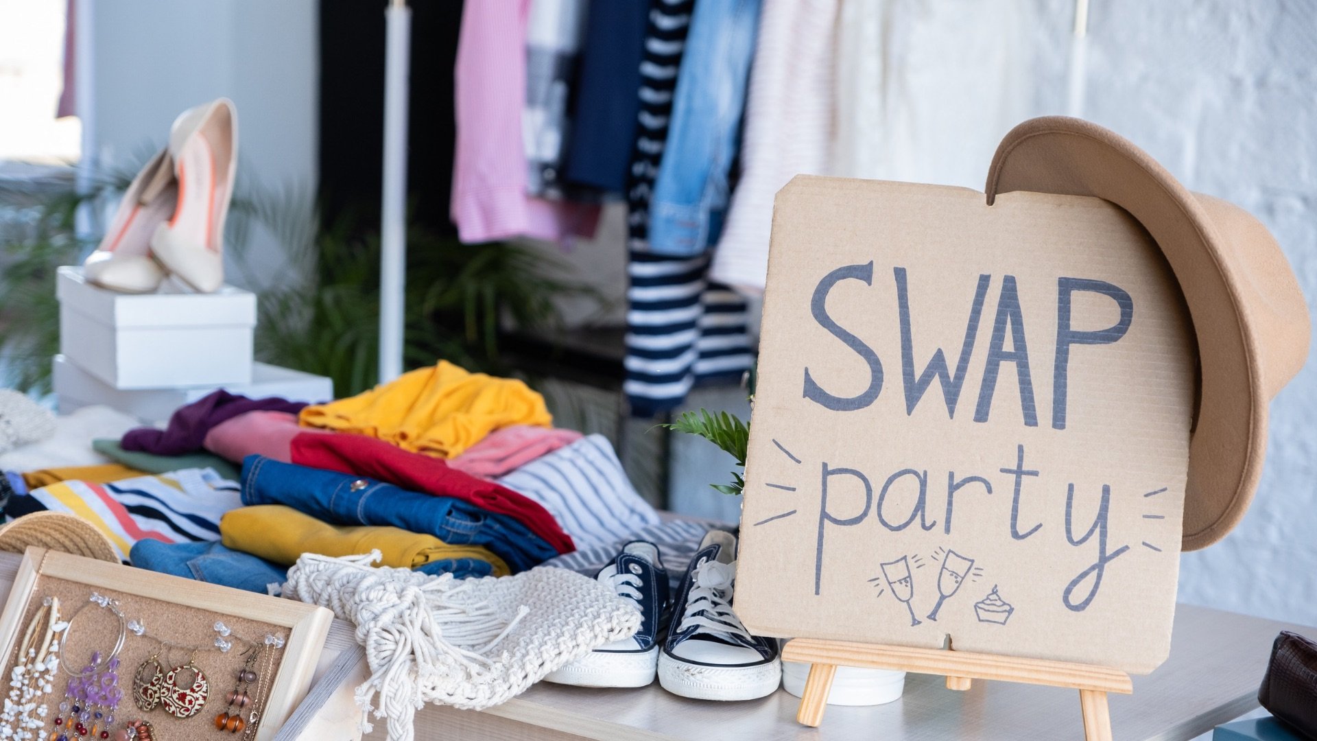 a picture of a clothing folded and stacked on a table. some heels are on a box and some dresses are hanging up in the background. One the table with the clothes there is a brown sign that reads clothing swap party. a hat is hanging on the sign.