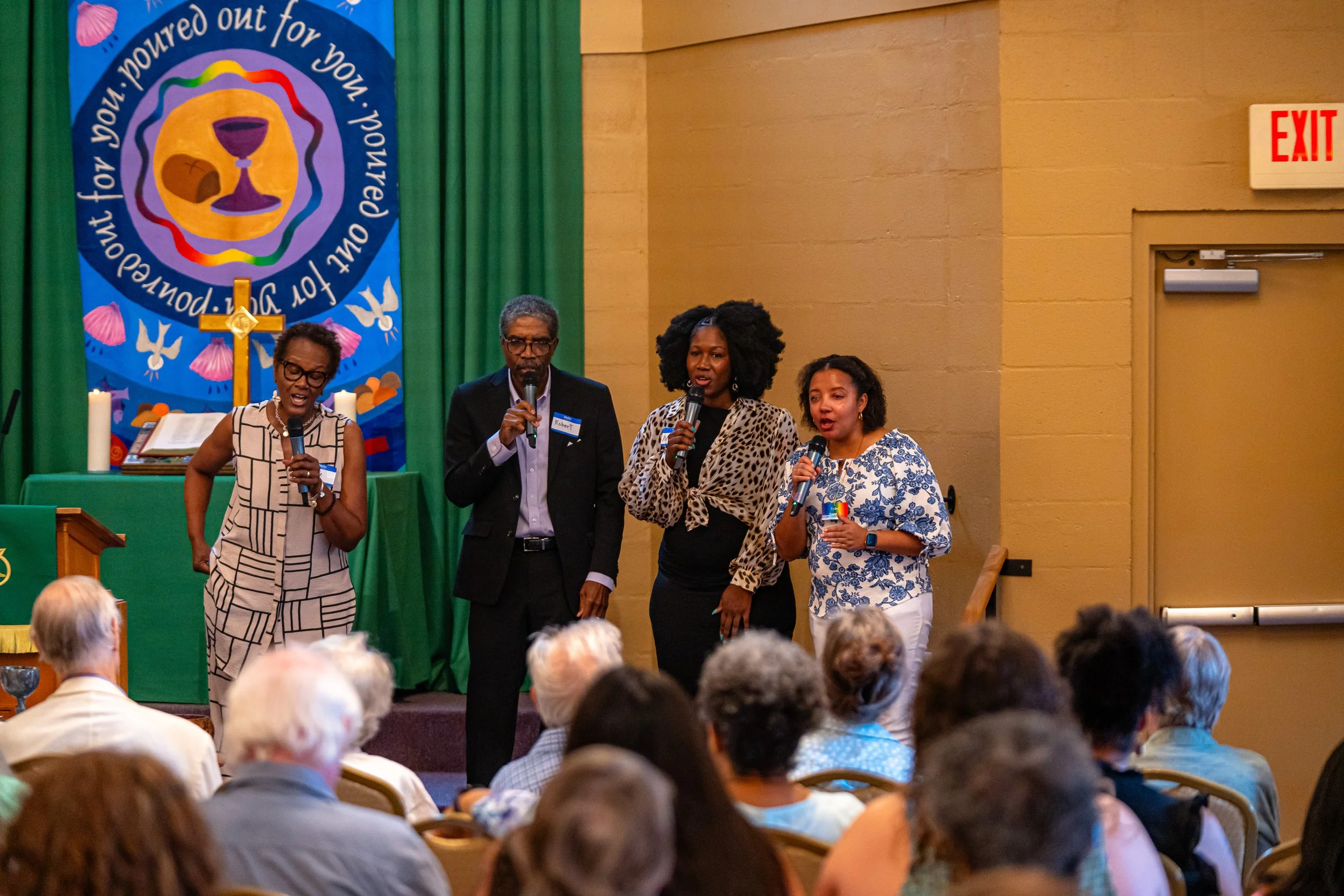 Four people are standing on a stage in front of an audience, each holding a microphone. The background features a blue banner with colorful symbols and text, and green curtains. Audience members are seated and facing the stage.