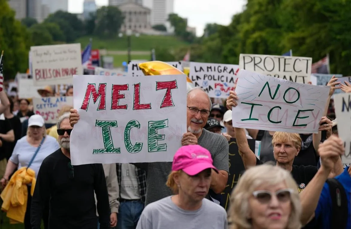 People protesting outdoors holding signs that say "MELT ICE" and "NO ICE" with a crowd and trees in the background.
