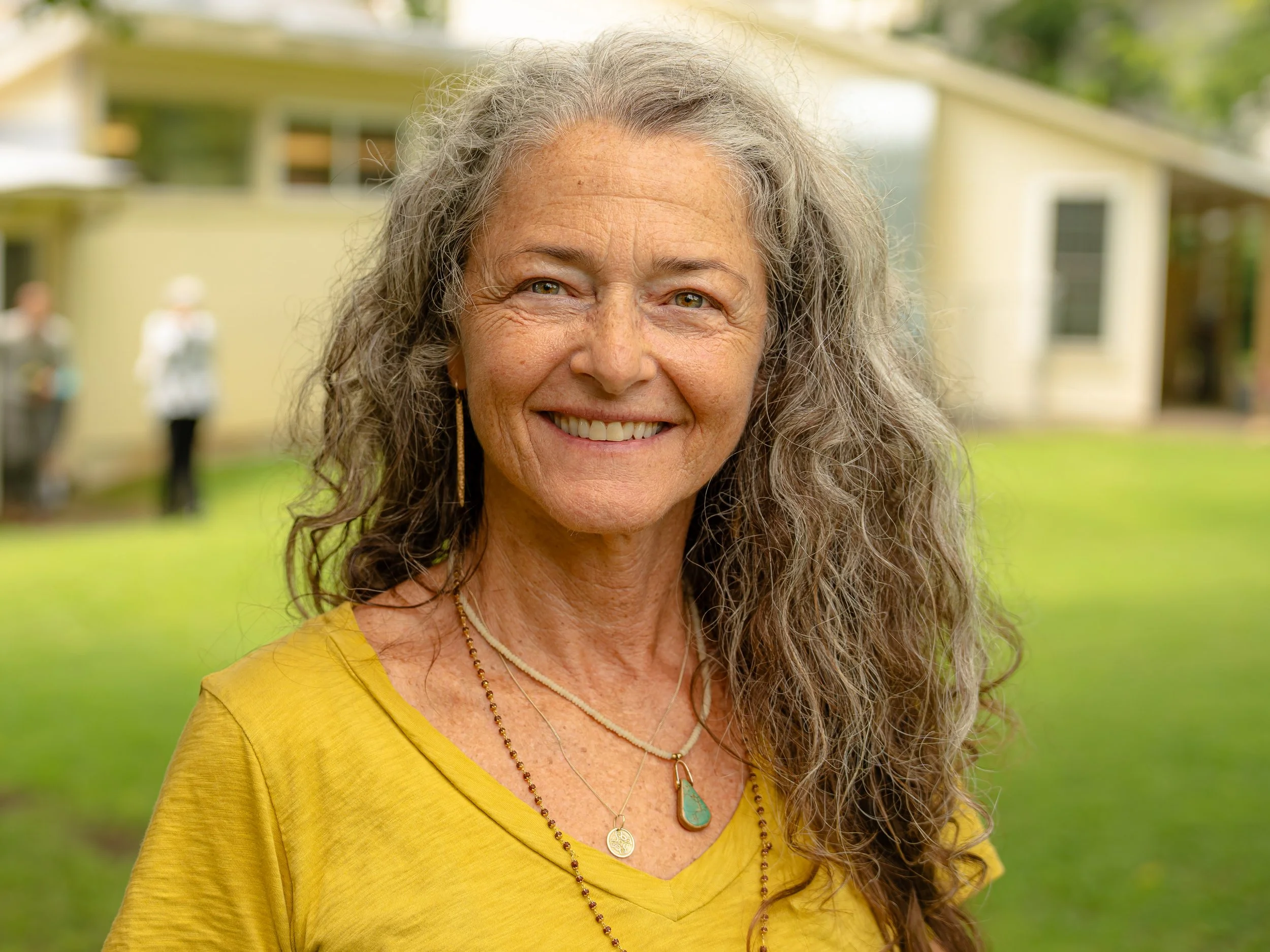 Close-up of an older woman with long, gray, curly hair smiling outdoors in front of a yellow house with green surroundings.