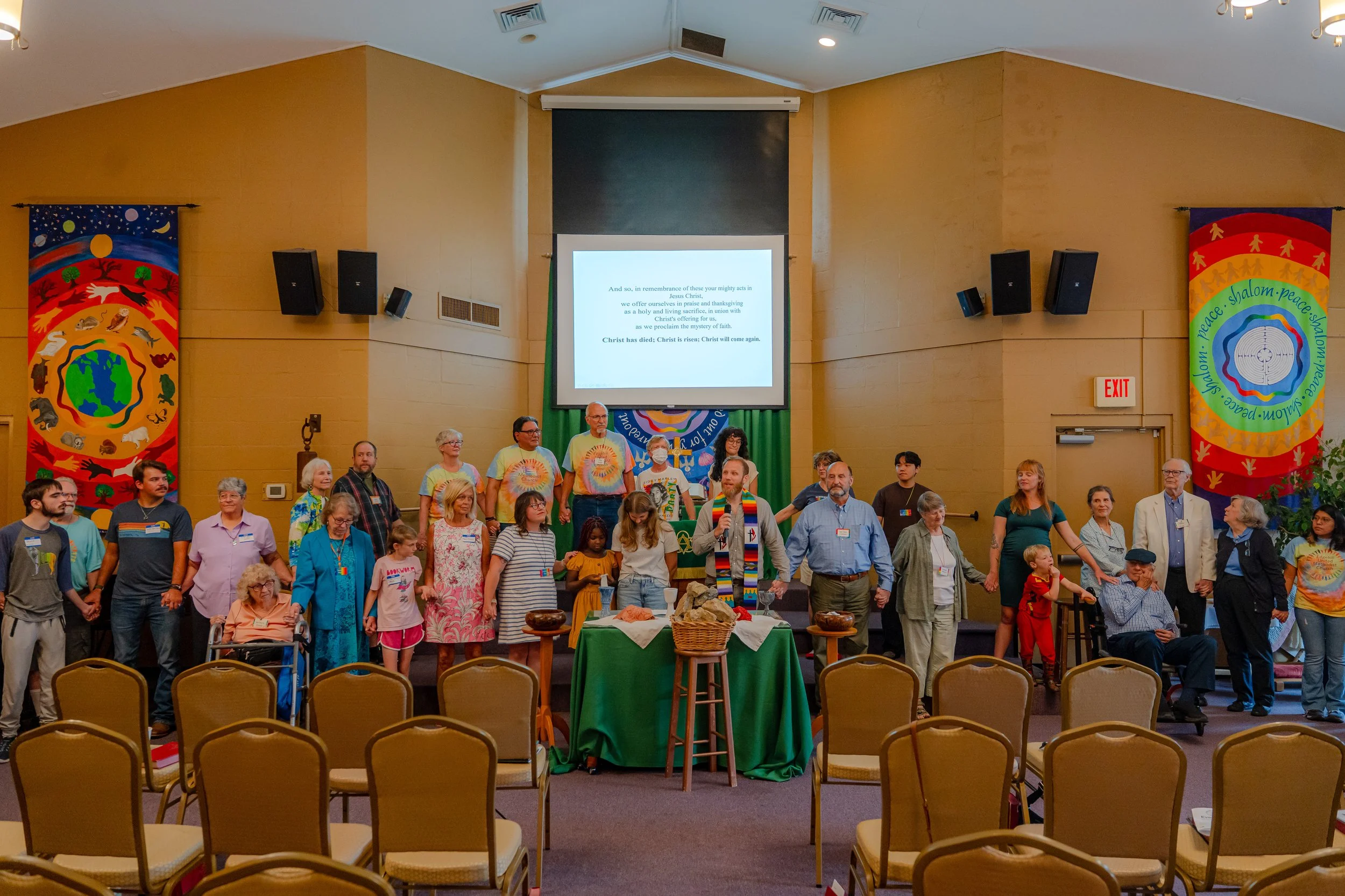 A diverse group of people gathered in a church or community hall for a service or event. The room is decorated with colorful banners with symbols of peace, shalom, and various animals around a globe. People are standing, some with hands raised or cla
