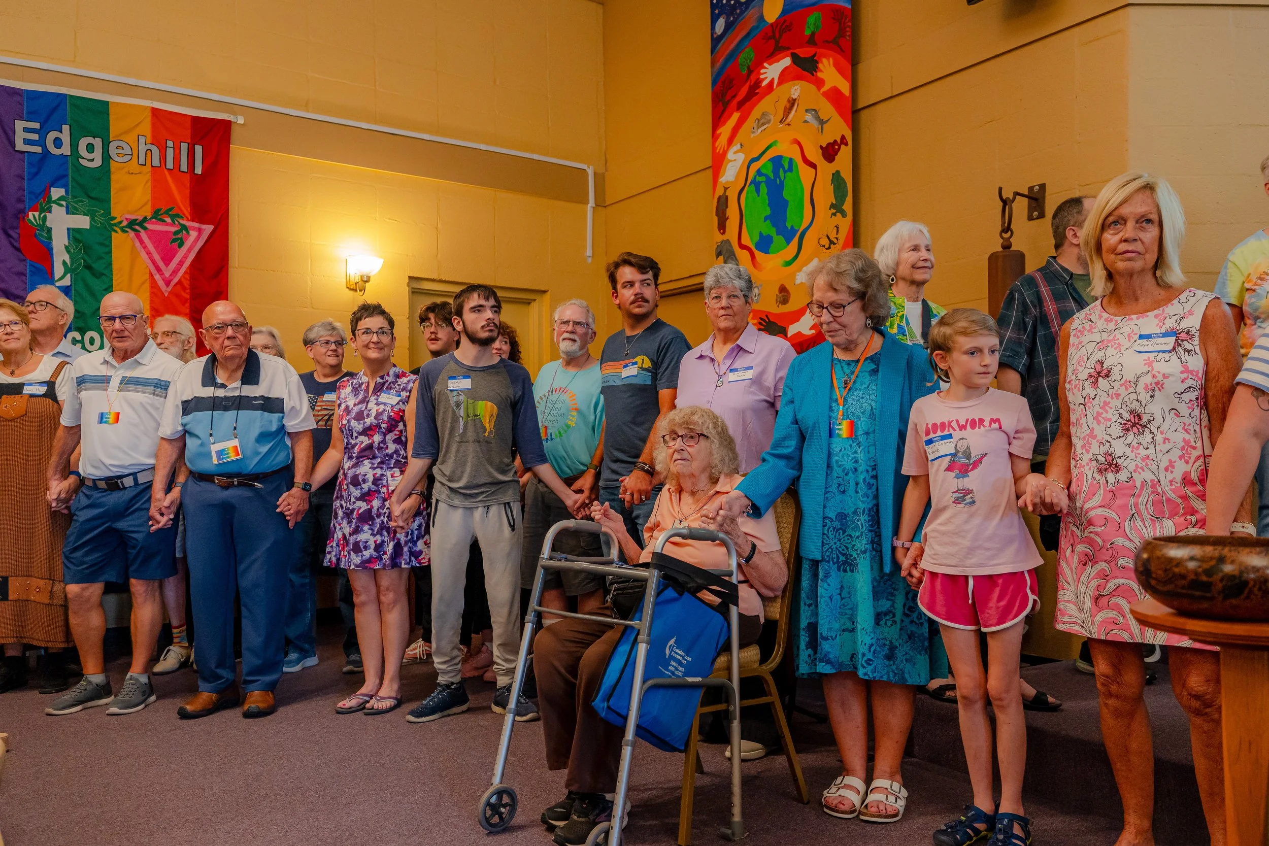 A diverse group of people, including elderly, middle-aged, and children, standing and sitting, gathered in a room decorated with colorful banners and artwork. Some are holding hands, and several are wearing name tags.