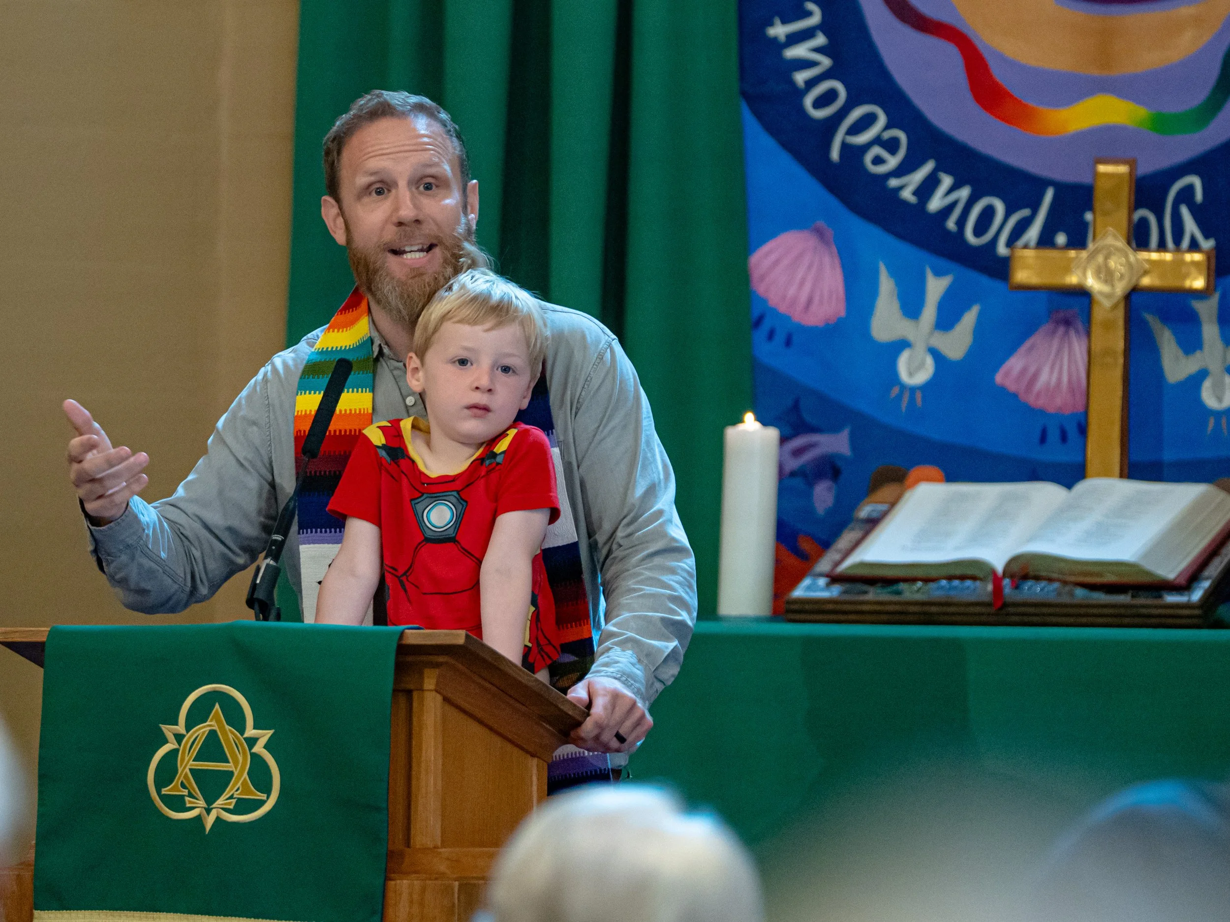 A man with a beard and a child with red hair are sitting at a wooden lectern during a ceremony. The man is wearing an autism awareness rainbow scarf and is speaking, while the child is sitting on his lap, looking forward. Behind them is a green curta