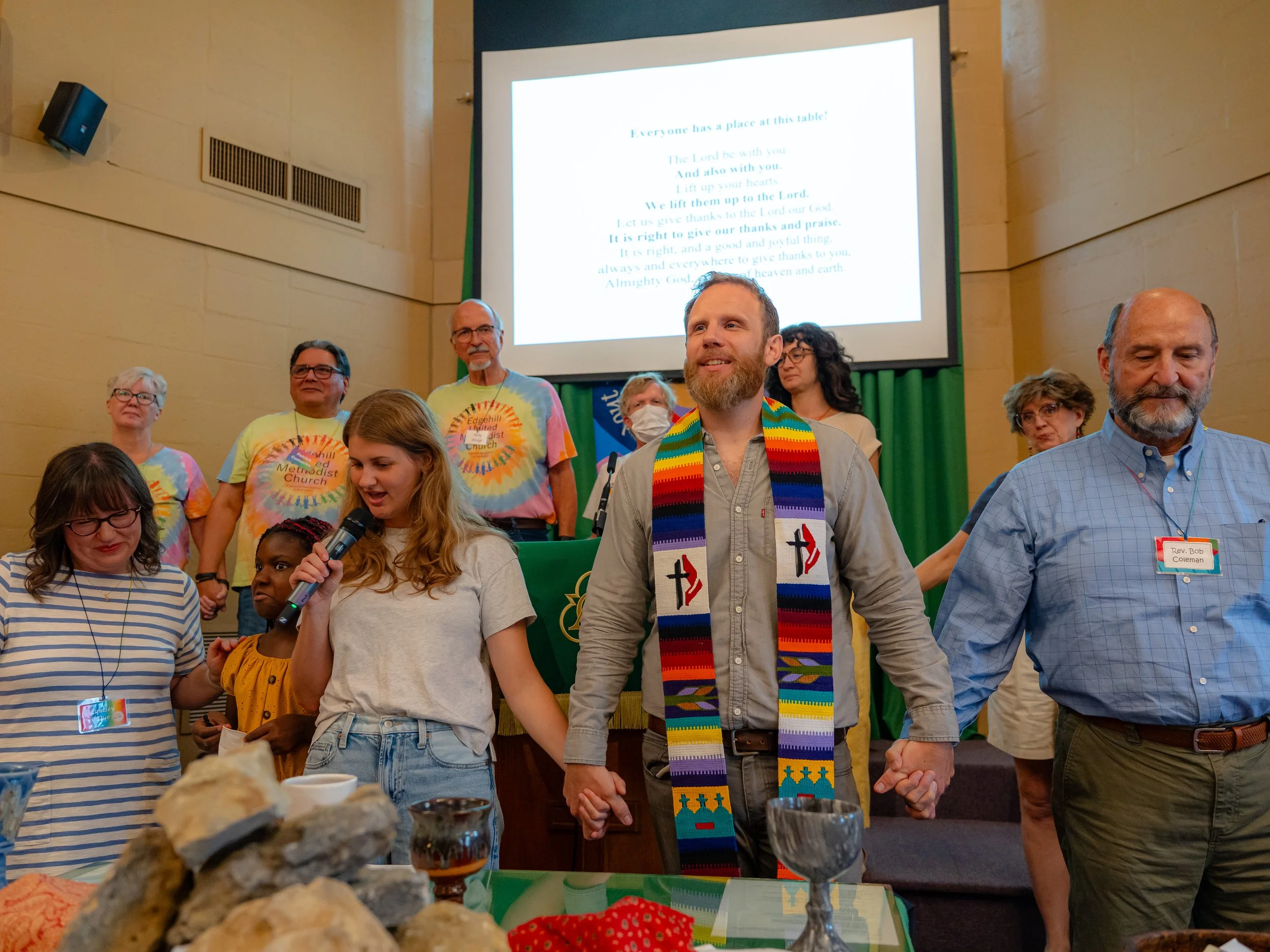 A group of diverse people standing together at a church event, some wearing colorful tie-dye shirts with the church's name, in front of a large screen displaying a religious quote. The setting appears to be a church hall with a table in the foregroun