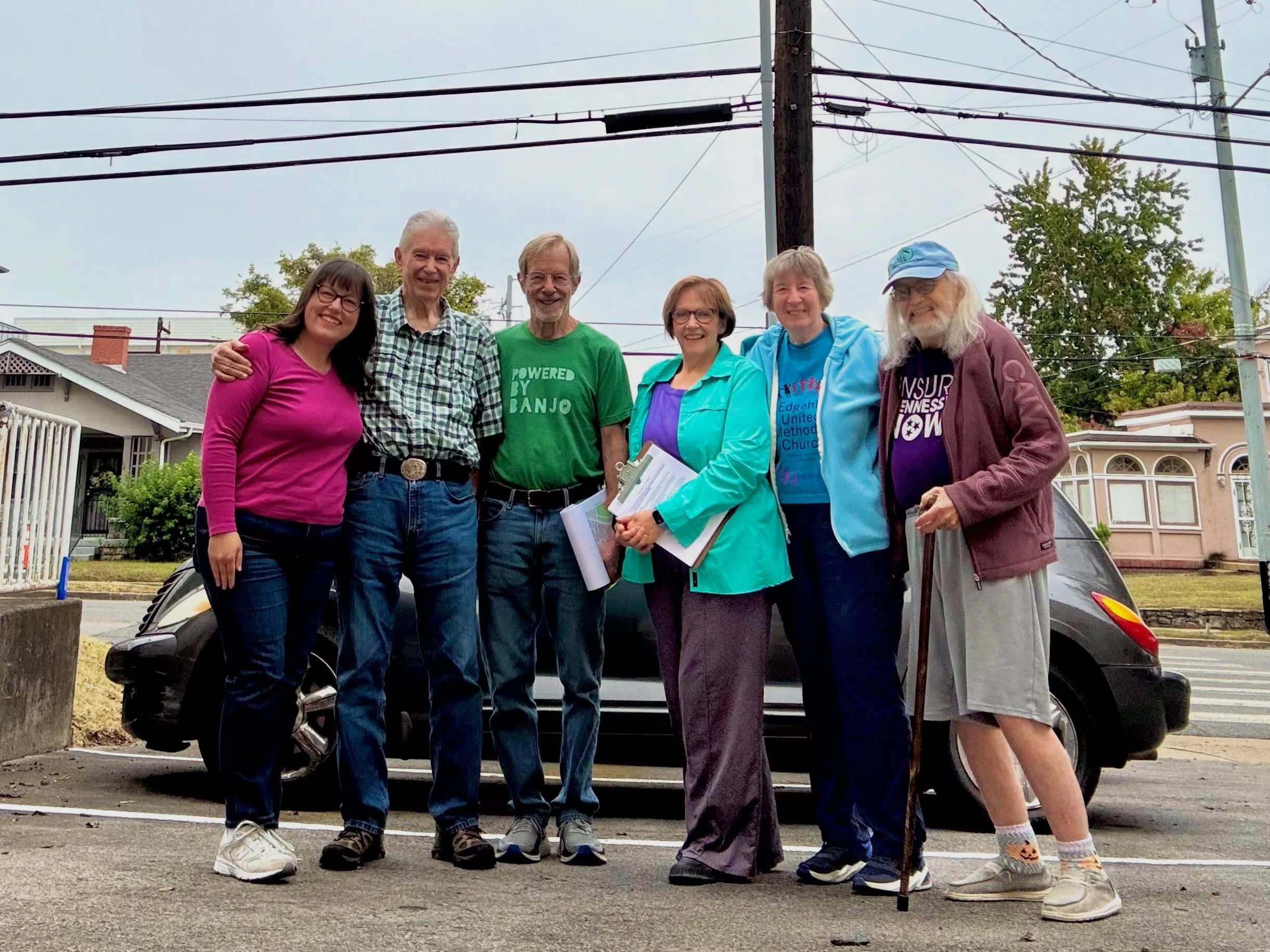 Group of six diverse people standing outdoors, smiling, in front of a car and electrical wires, under an overcast sky.