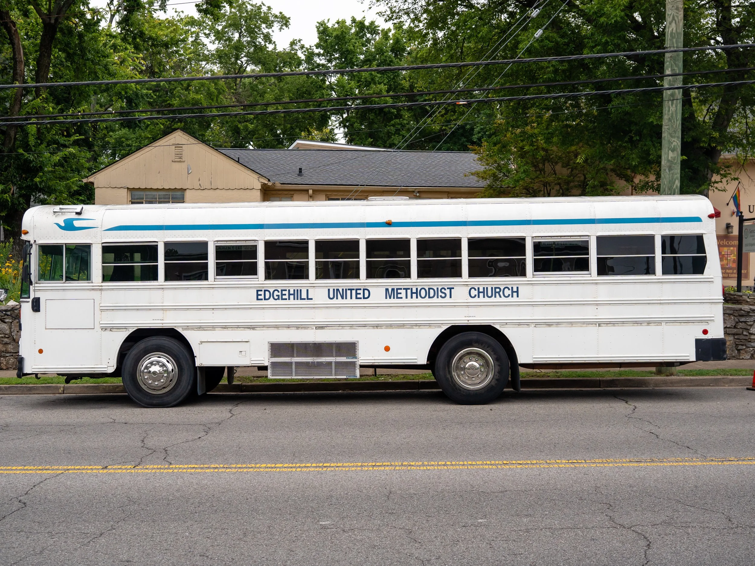 A white bus with blue stripes parked on a street in front of a house with a gray roof and beige siding. The bus has the words "Edgehill United Methodist Church" on its side and a blue wave symbol near the front.