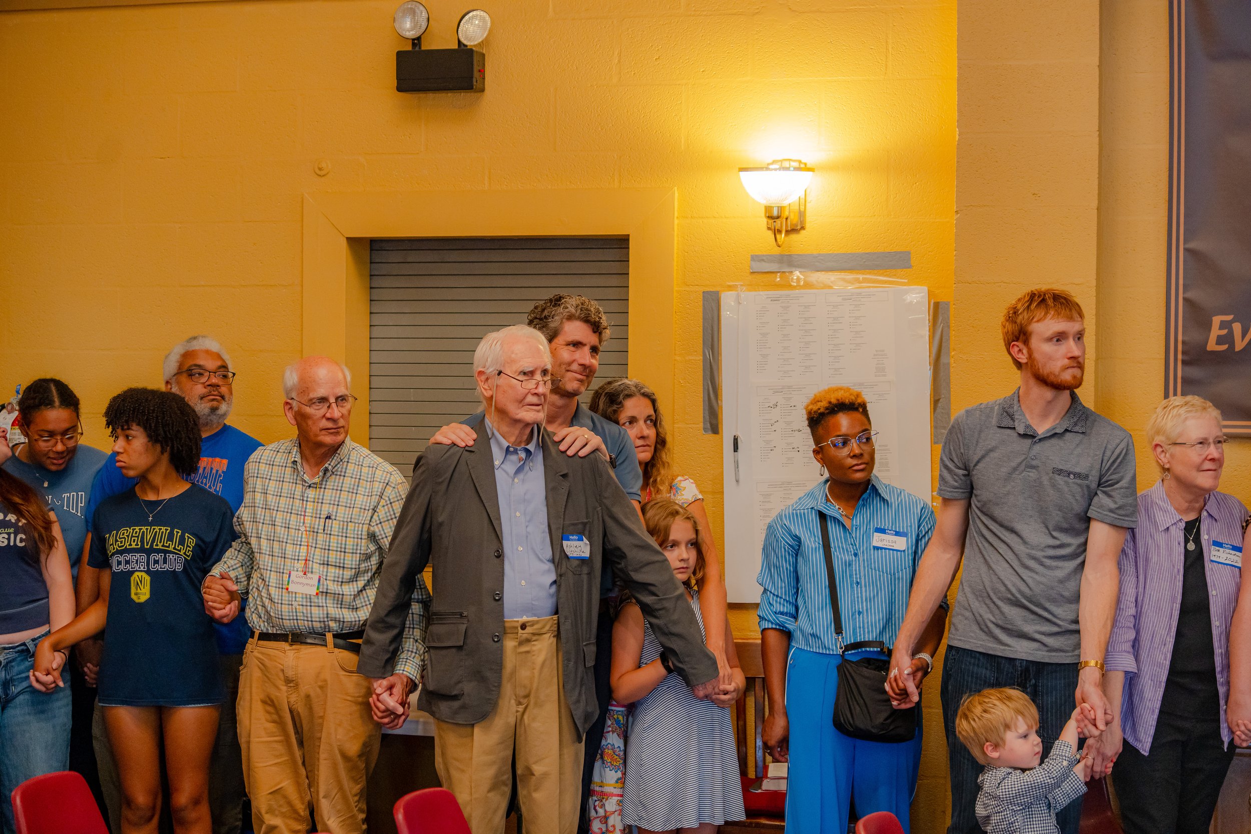 Group of diverse people standing together indoors, holding hands, in a prayer circle during an event.