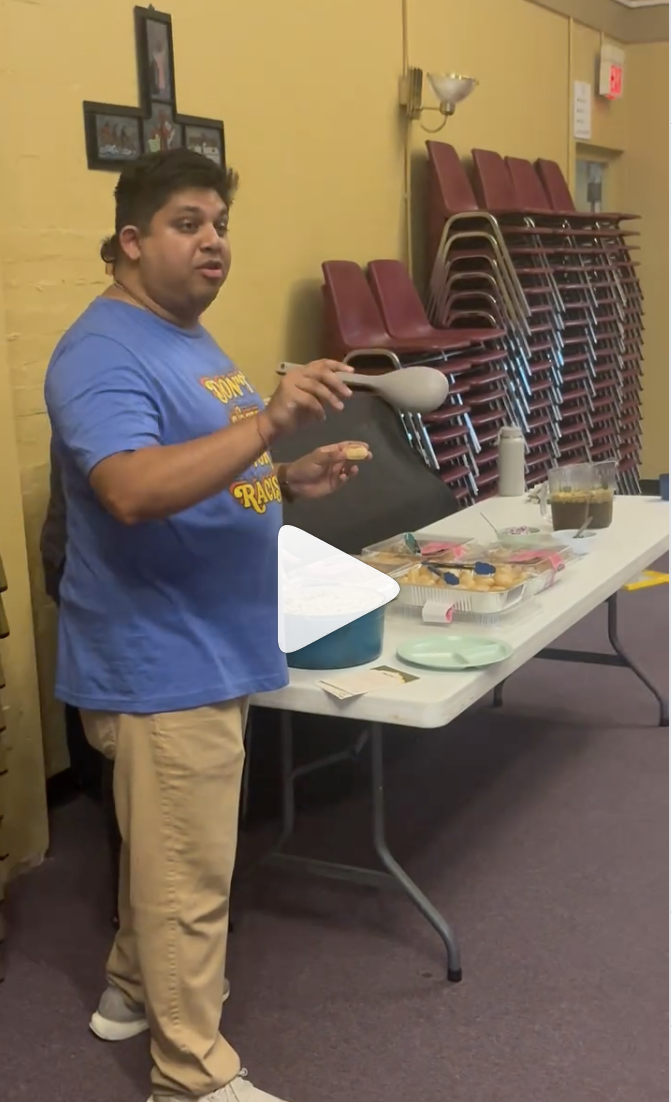 A man in a blue T-shirt and beige pants stands at a table with snacks and drinks, holding a popcorn scoop. Behind him is a yellow wall with stacked chairs and a cross-shaped photo frame.