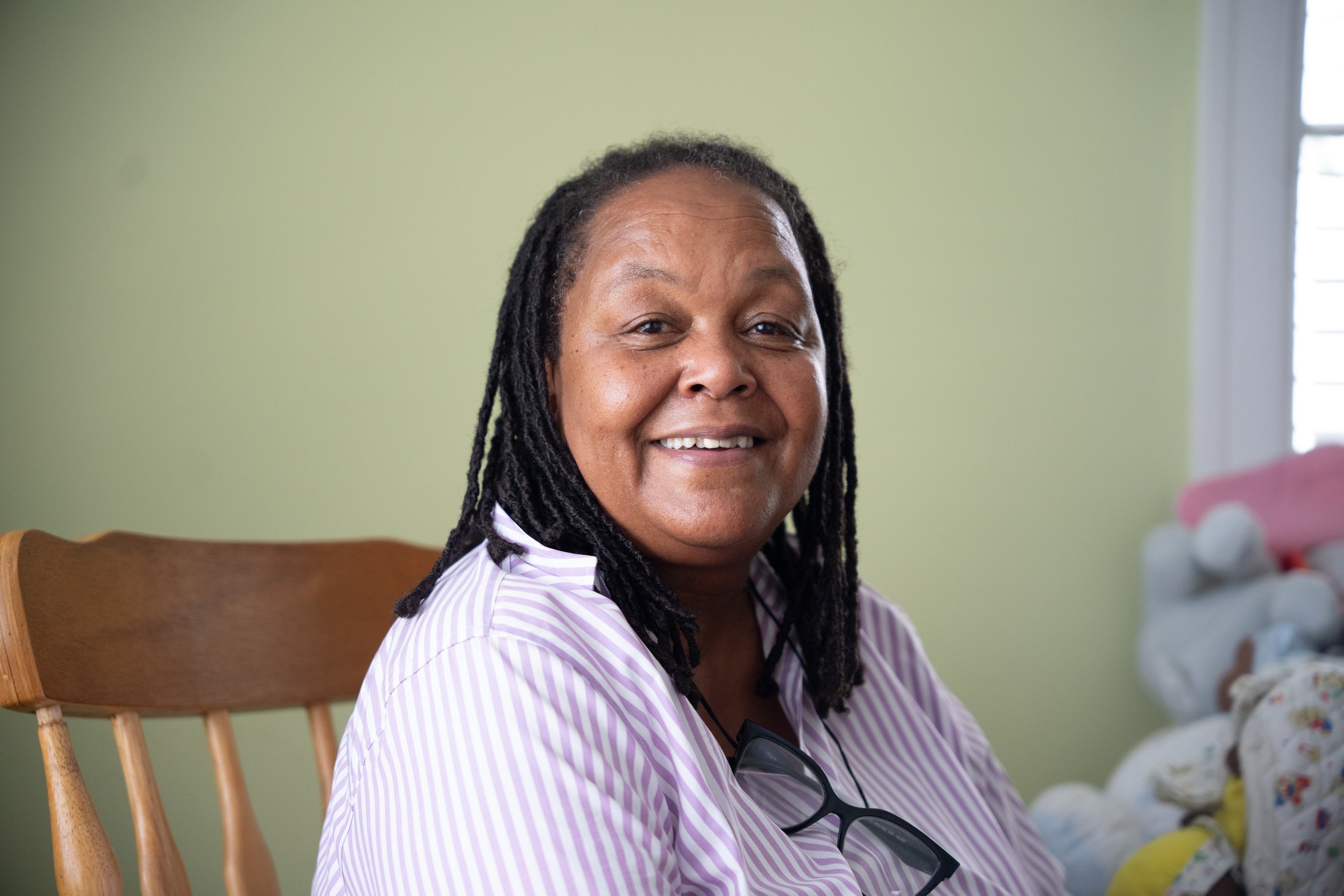 A smiling woman with dreadlocks sitting on a wooden chair indoors, wearing a striped shirt and glasses hanging from her neck, with a baby in a floral outfit nearby.