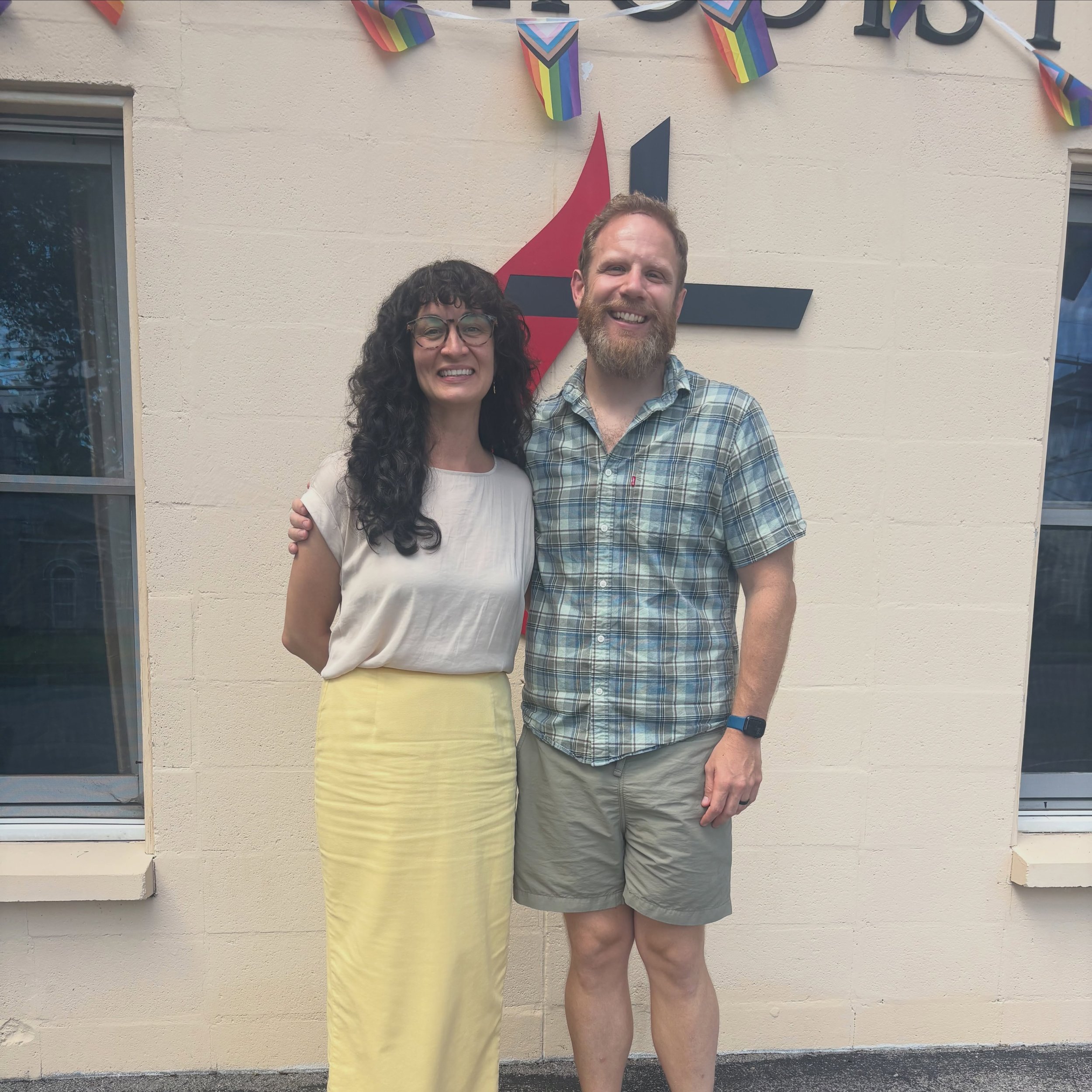 Two people standing close to each other in front of a wall with rainbow pride decorations. One person is a woman with curly dark hair, glasses, and a white top, and the other is a man with a beard, short hair, wearing a plaid shirt and shorts. They are smiling.