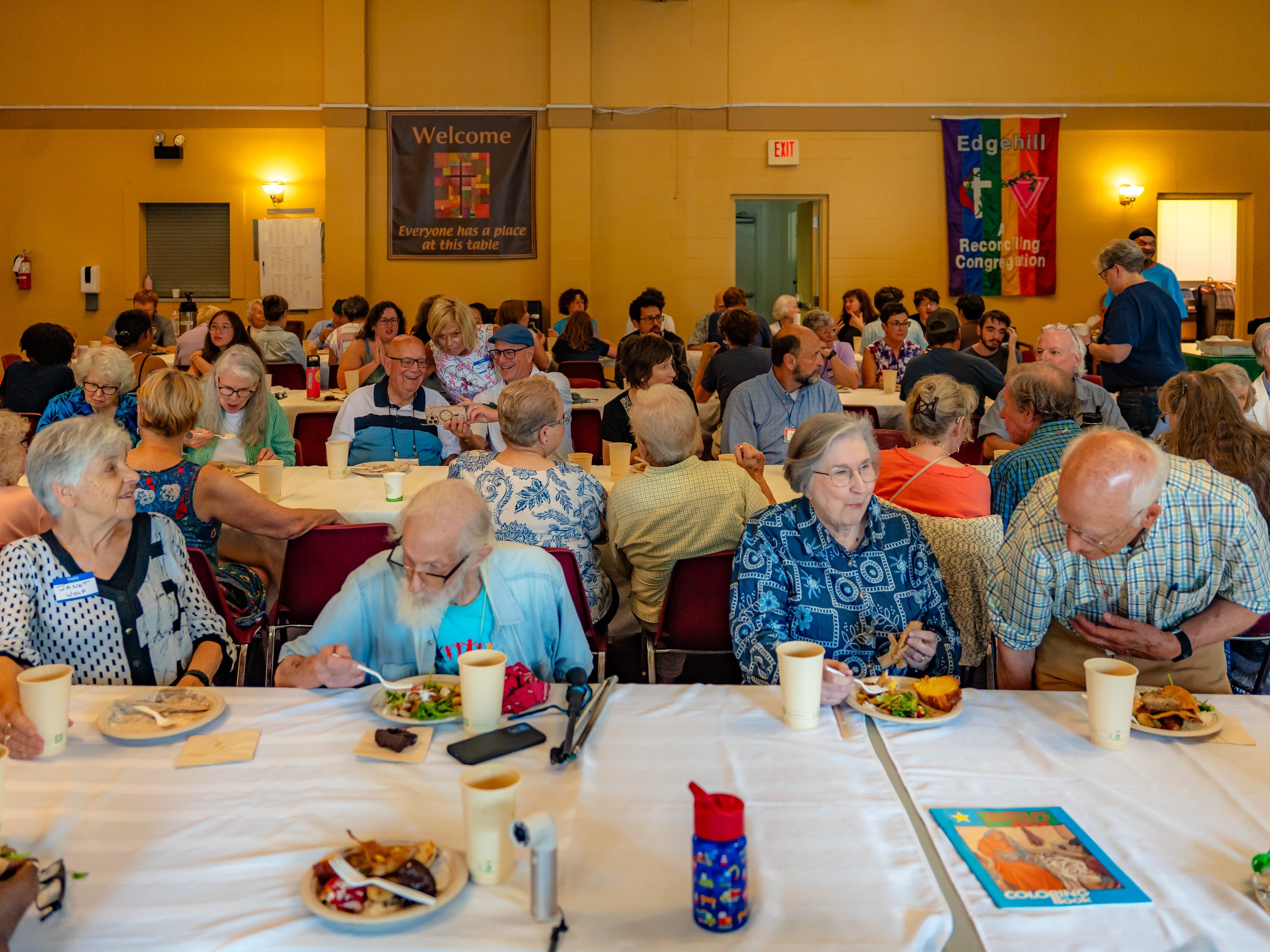 A large group of people sitting at tables in a banquet hall, enjoying a meal and socializing. The walls have welcoming signs and colorful banners; the atmosphere appears cheerful and lively.