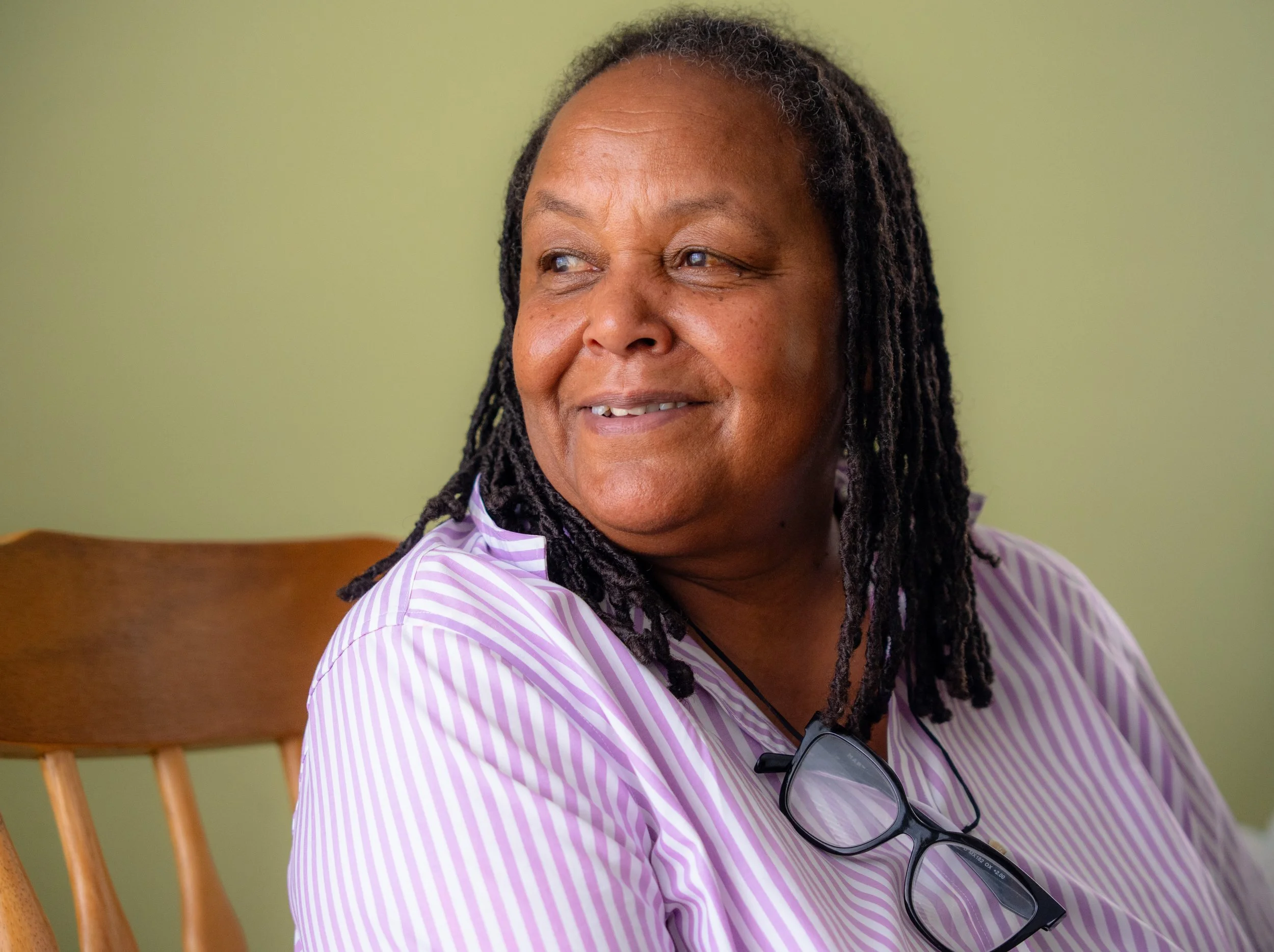 A middle-aged woman with dark skin and dreadlocks wearing a purple and white striped shirt, smiling and looking to her left, sitting on a wooden chair against a light green wall. Sunglasses are hanging from her shirt.