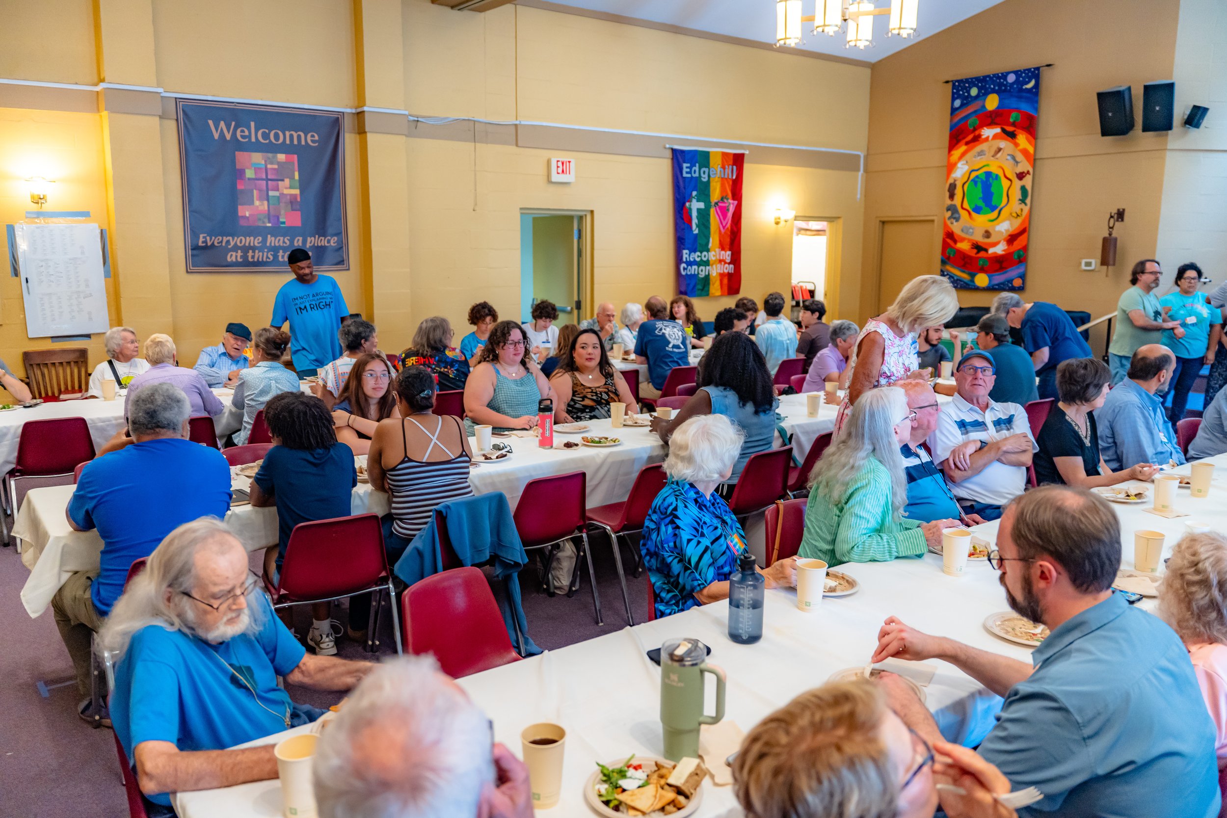 A large group of diverse people gathering in a hall for a meal, sitting at tables with food, on a sunny day. The hall has yellow walls, colorful banners, and paintings, with ceiling lights and speakers.