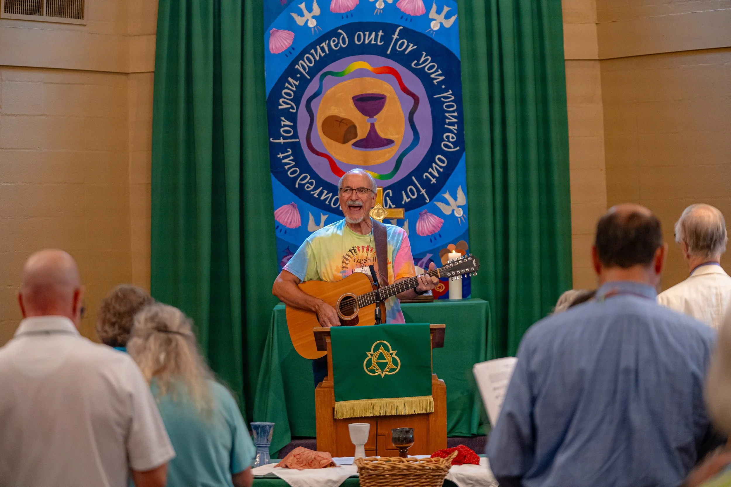 A man with glasses and a gray beard wearing a colorful tie-dye shirt with a guitar, singing or speaking in front of an audience. Behind him is a large banner with religious symbols and the text "poured out for you." The audience is seated at tables, 