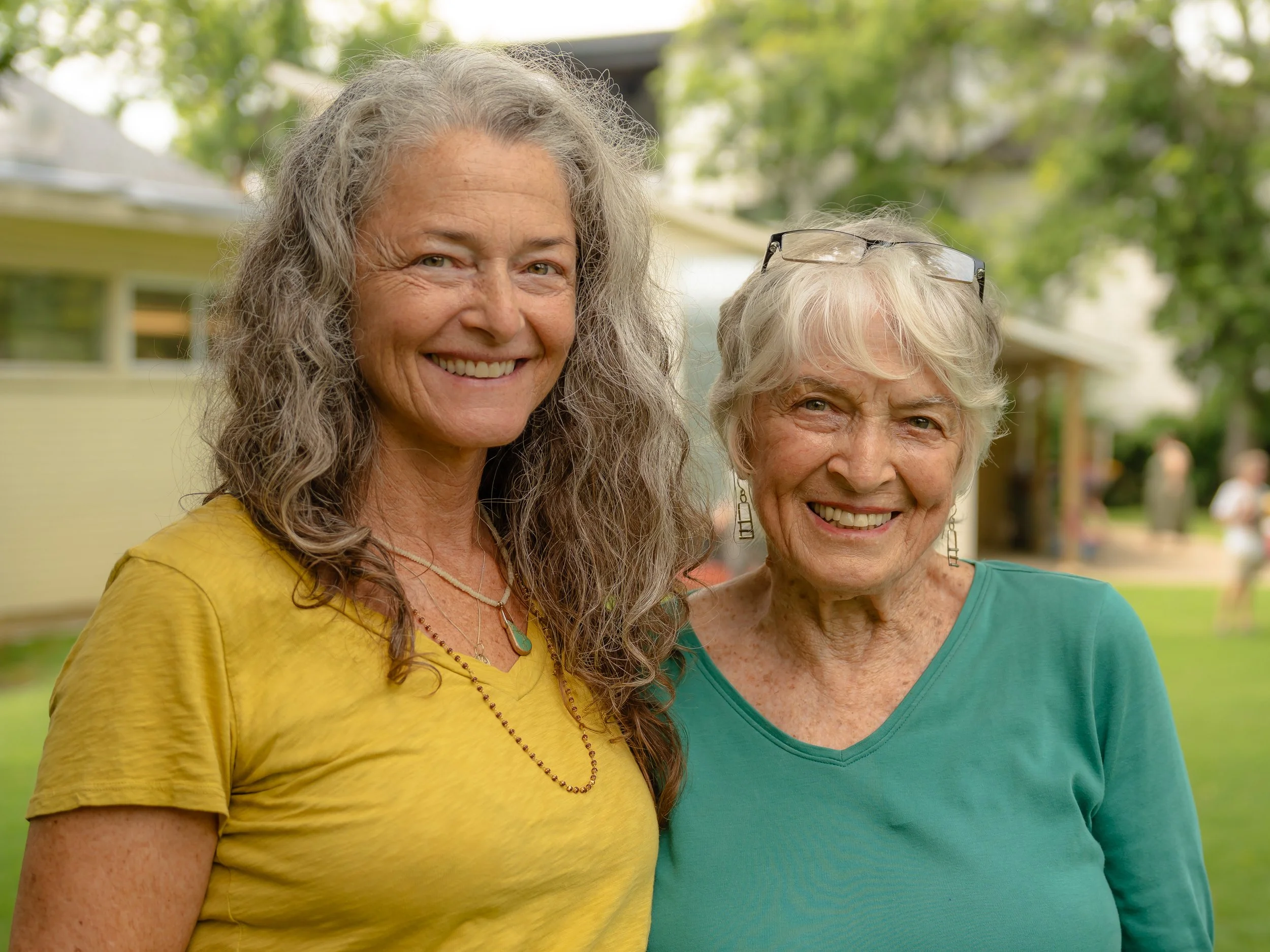 Two smiling elderly women with gray hair, one wearing glasses on her head, standing outdoors in front of a green, leafy background.