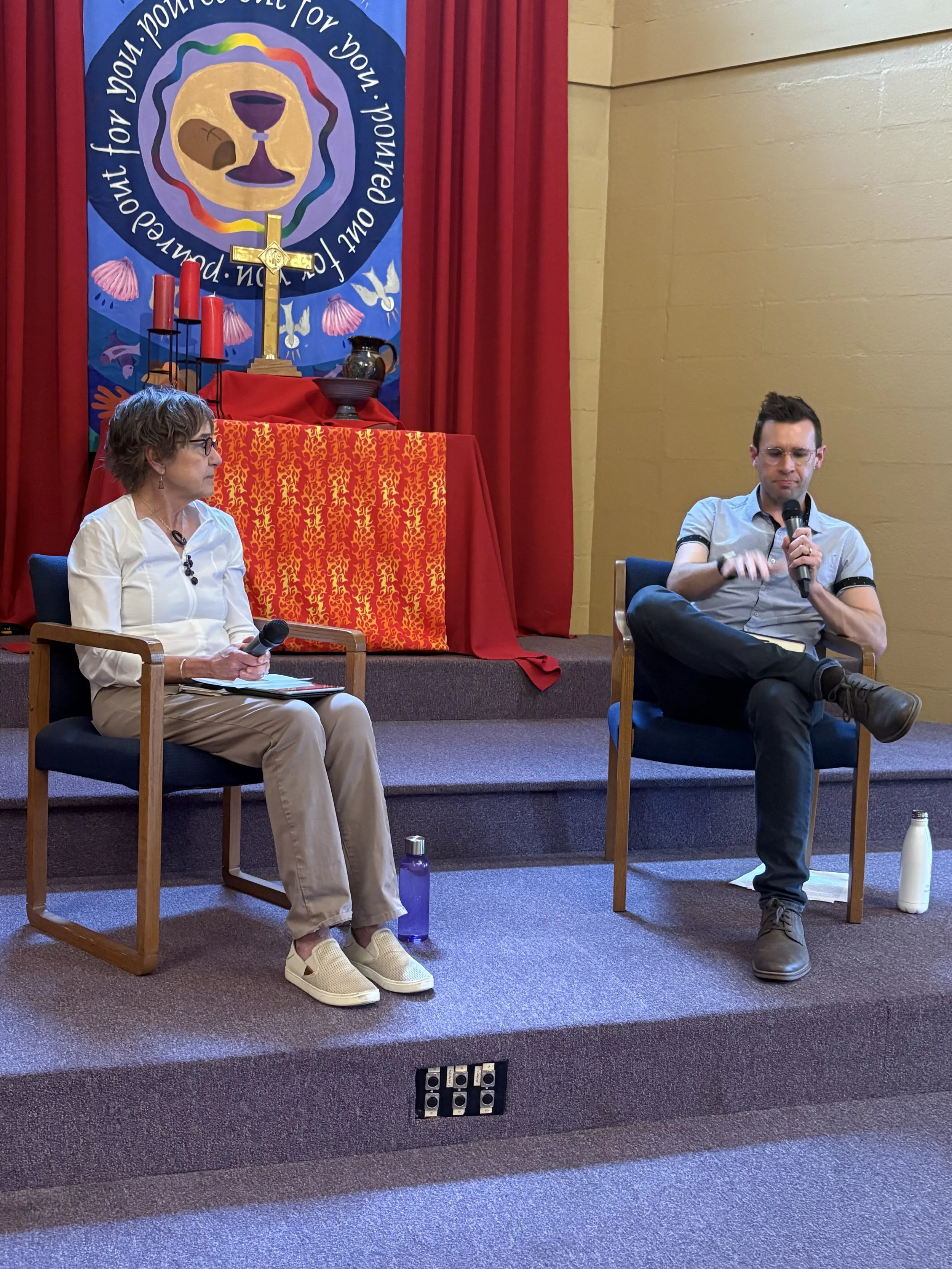 A woman and a man sitting on chairs on a stage during a panel discussion, with a religious altar in the background displaying a cross, candles, and a banner with religious symbols and text.