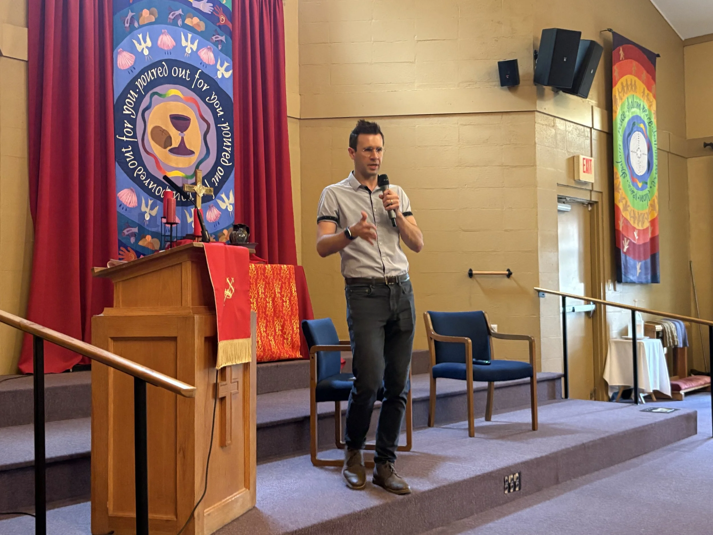 A man is speaking into a microphone on a stage with red curtains and religious banners hanging behind him. The banners have symbols and text related to a religious or church setting.