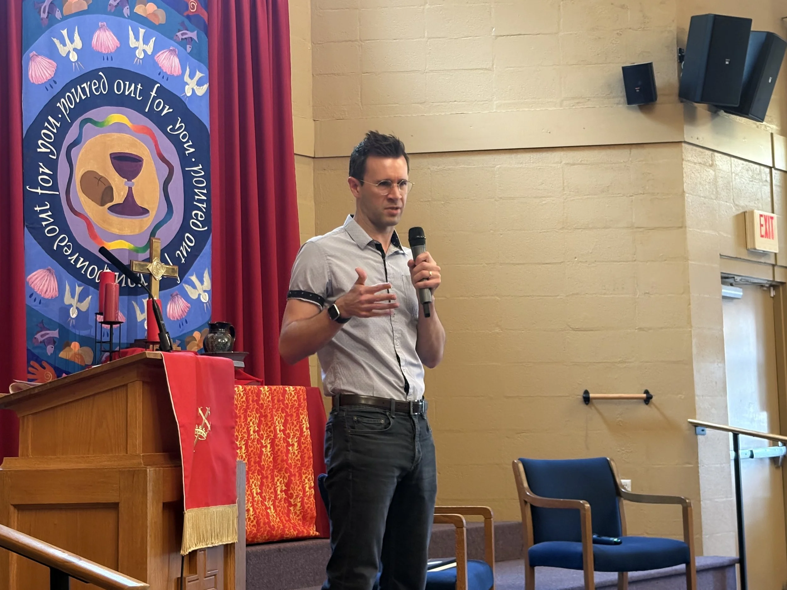 A man in a light gray shirt and dark glasses is speaking into a microphone inside a room with beige walls and a red curtain. Behind him is a colorful religious banner with a chalice, bread, and symbols, along with pink dove illustrations and the phra