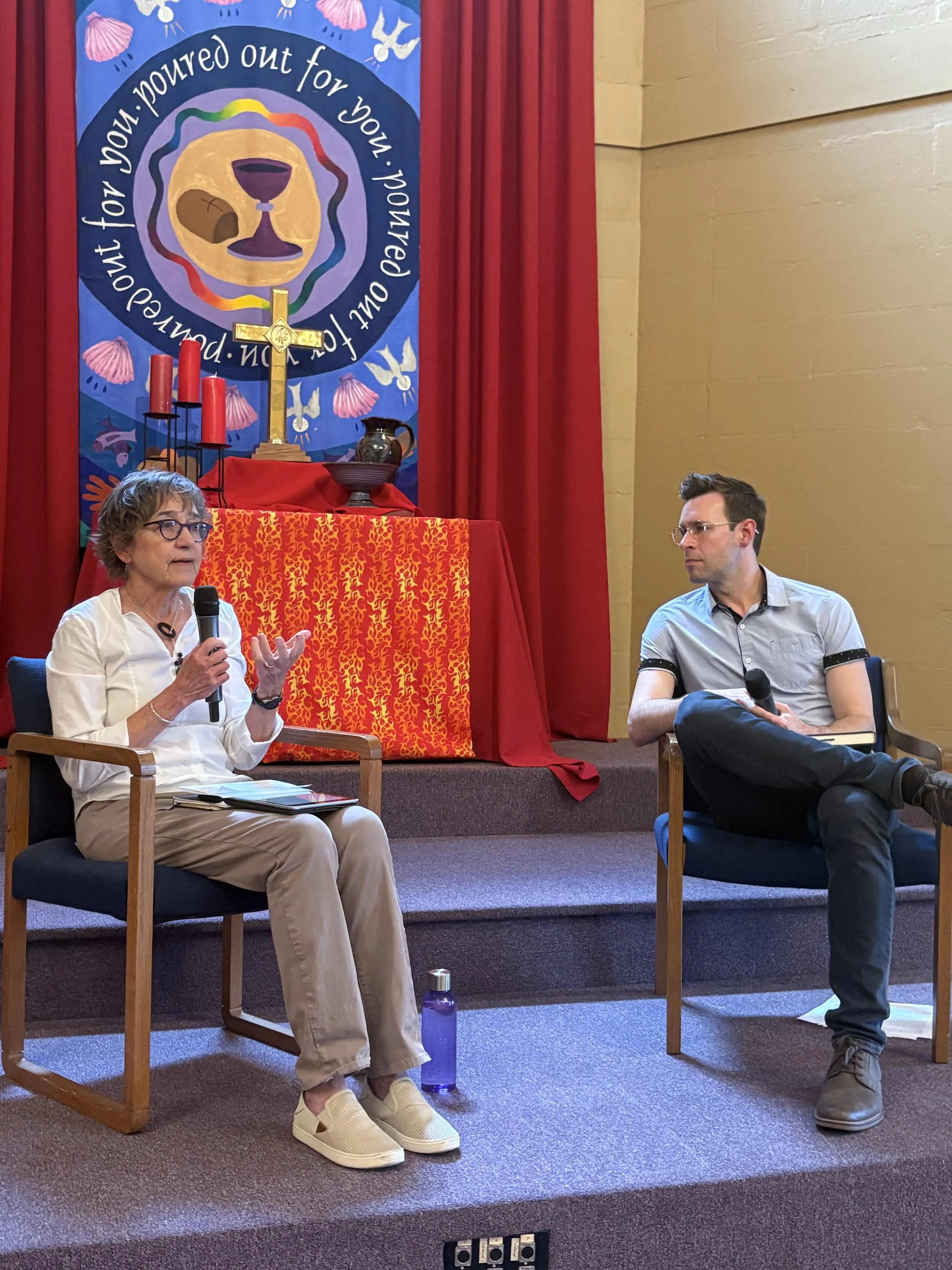 Two individuals seated on stage in conversation, one woman speaking with a microphone and one man listening, with a decorated backdrop featuring religious symbols, candles, and cloth.