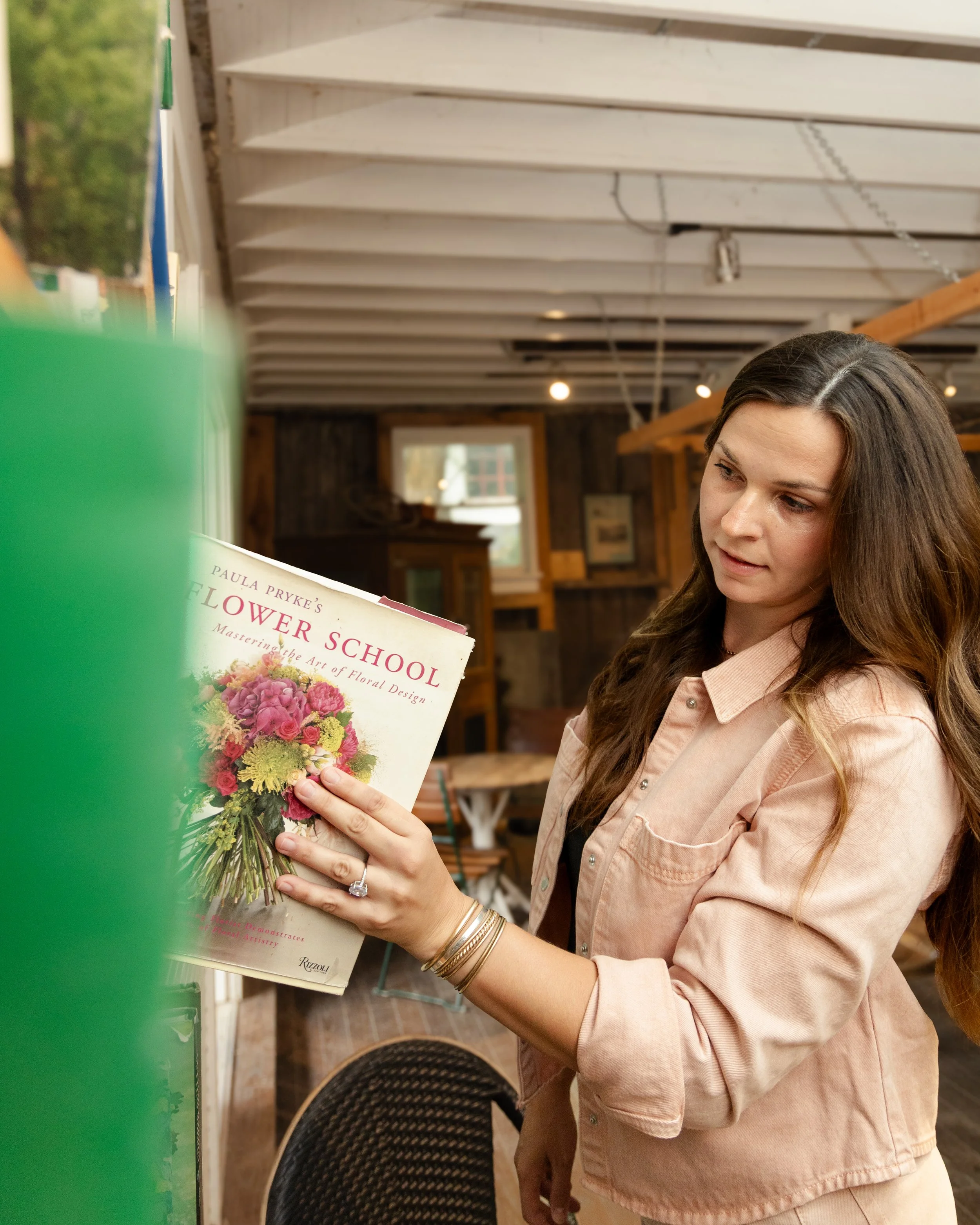 A woman with long brown hair looking at a book about floral design, holding it with both hands. She is wearing a light pink or beige button-up shirt and has multiple bangles on her wrists. The background shows a rustic indoor space with wooden beams, bookshelves, and a window.
