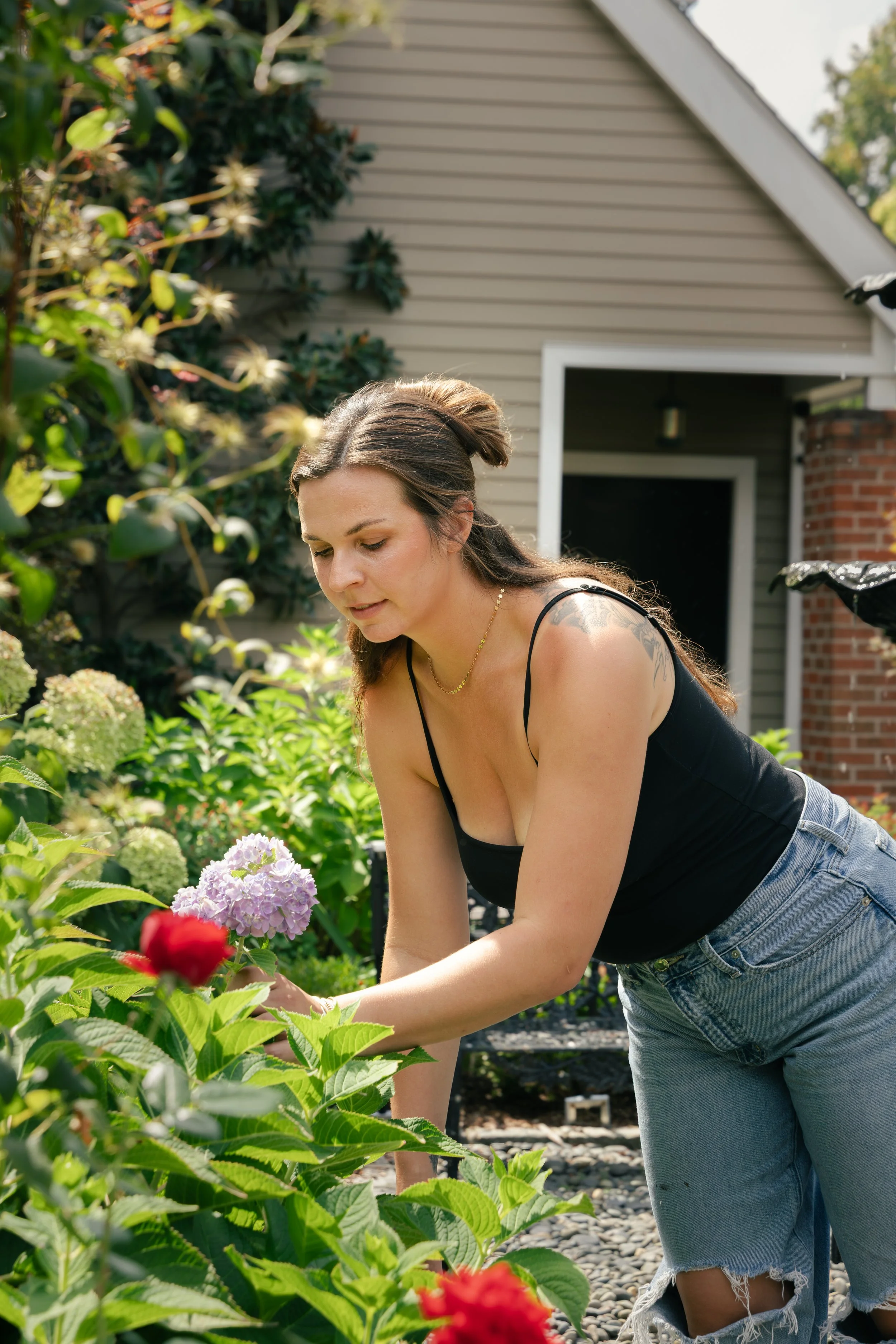 A woman wearing a black tank top and ripped jeans, with long brown hair, is gardening outside, surrounded by colorful flowers and greenery, in front of a house with beige siding and a brick column.