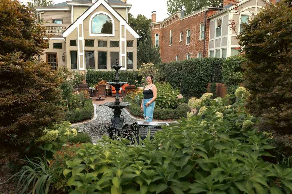 A woman standing in a lush backyard garden with a fountain in the center, surrounded by flowering bushes and greenery, with houses in the background.
