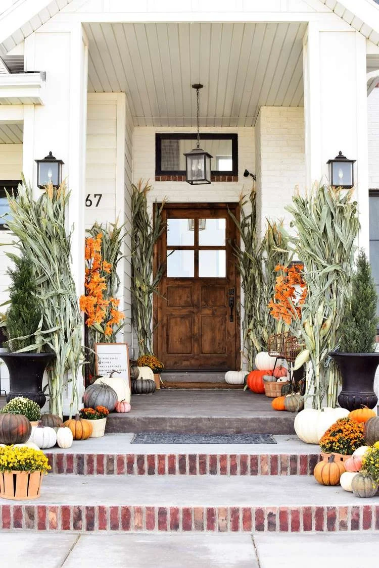 Festive autumn porch decorated with pumpkins, gourds, corn stalks, and potted flowers, featuring a wooden front door and black lantern-style lights.