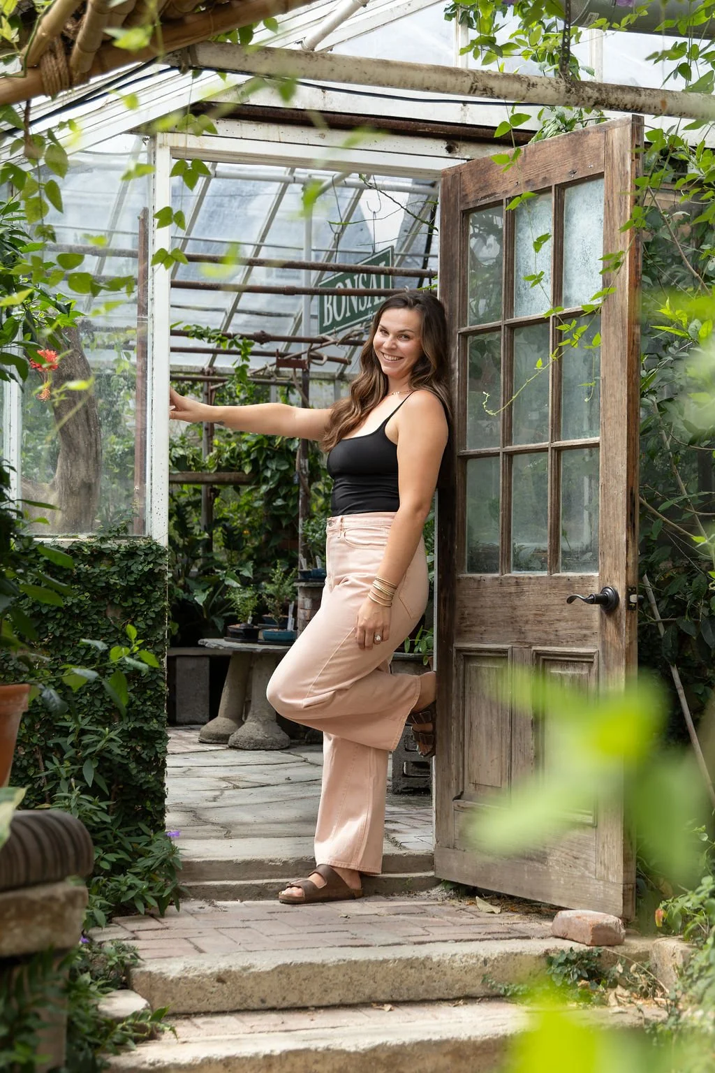 A young woman with brown hair, wearing a black tank top and light pink pants, smiling while standing in the doorway of a greenhouse surrounded by green plants.